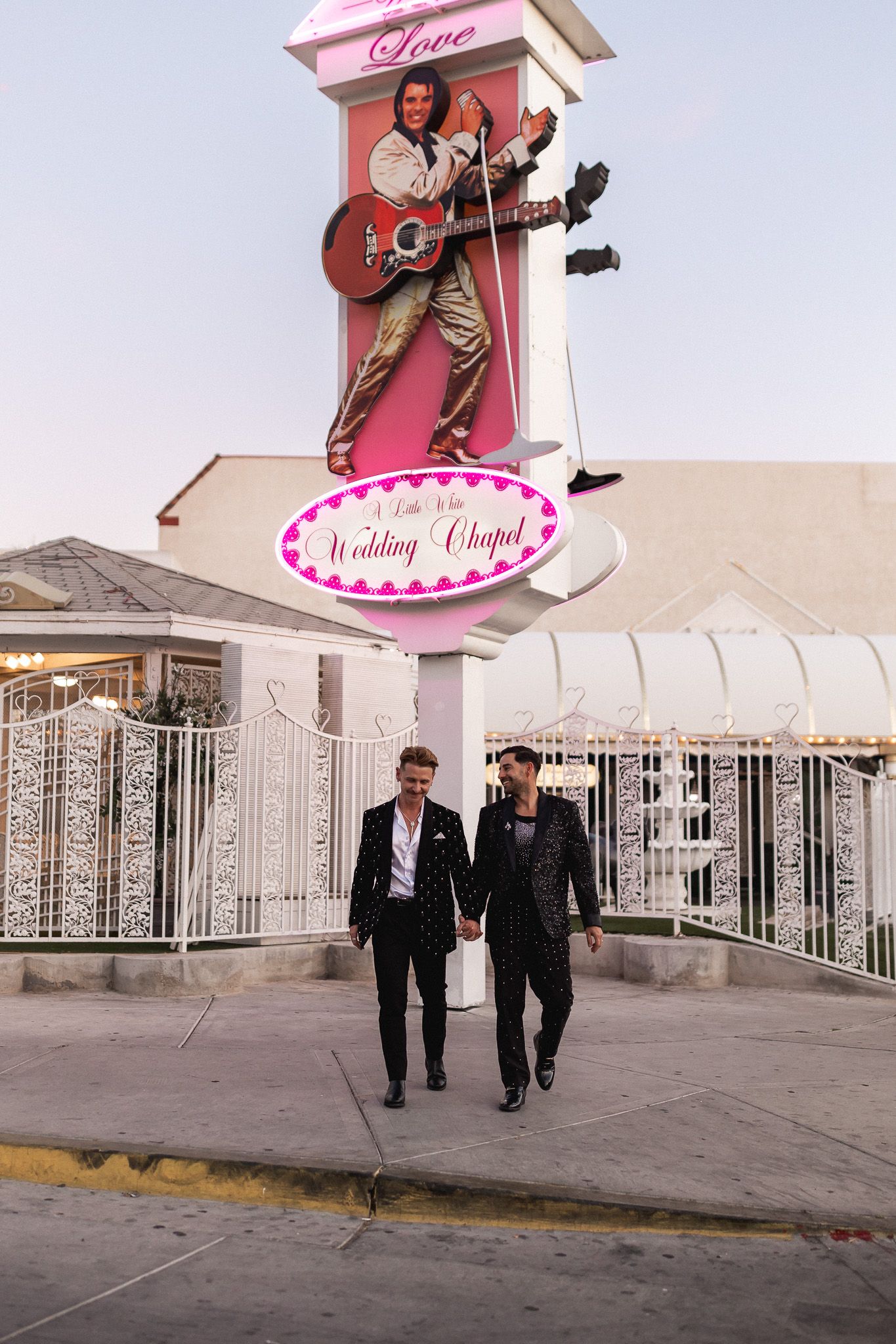 Two grooms hold hands walking in front of A Little White Wedding Chapel Elvis sign in Las Vegas.