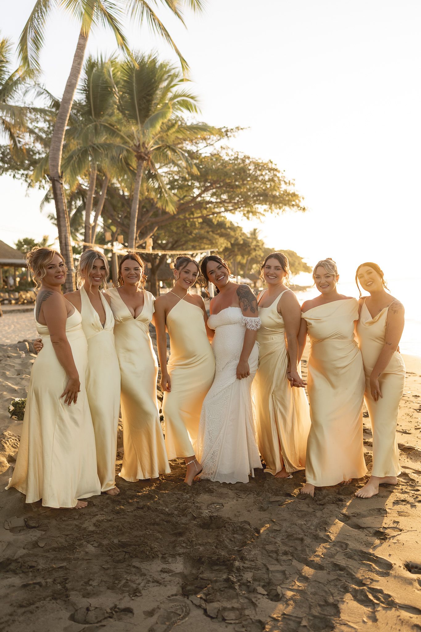 Bride and bridesmaids in cream gowns pose on sandy beach at golden sunset with palm trees.