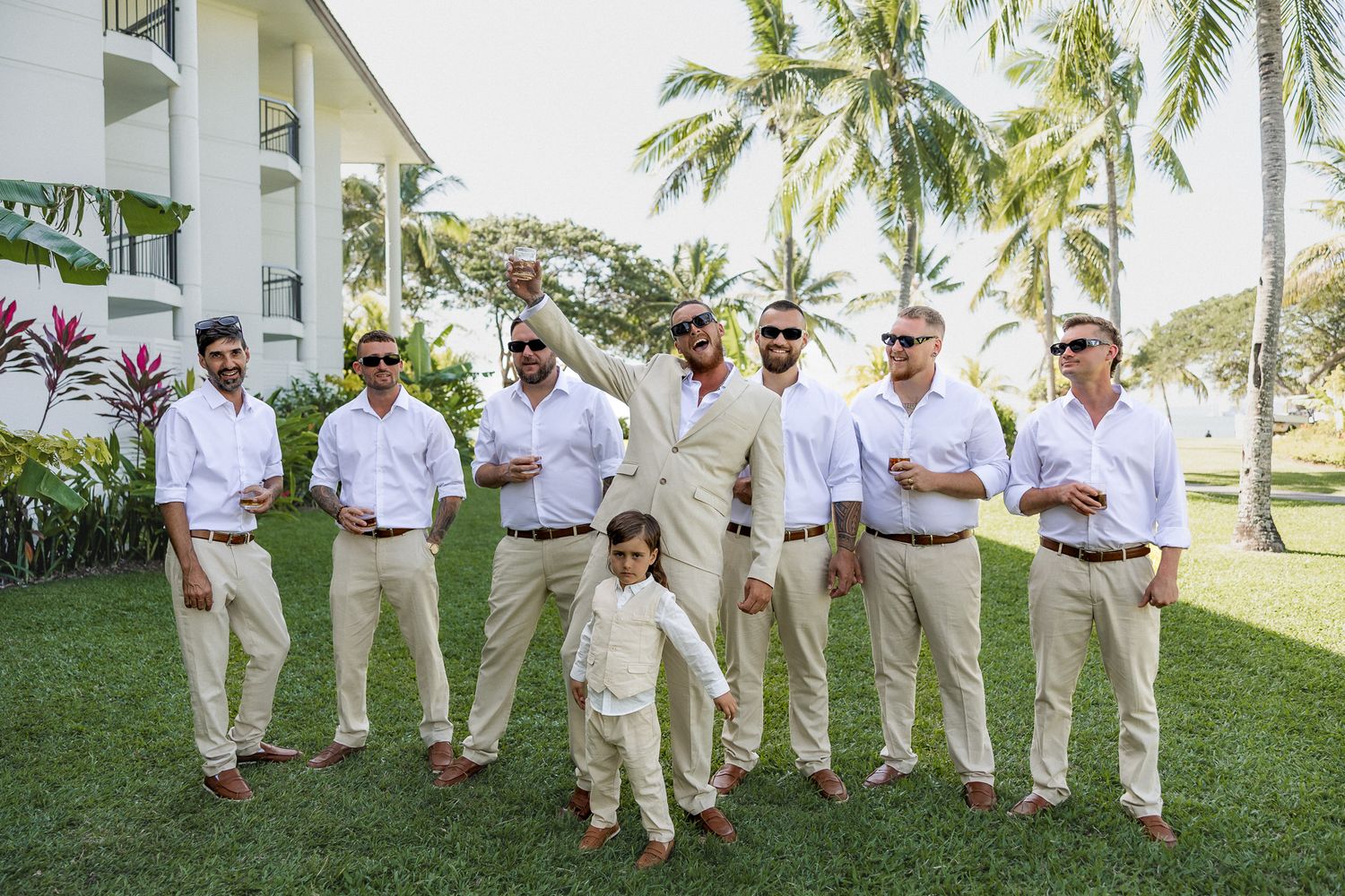Groomsmen in white shirts and khaki pants pose outdoors with palm trees at a tropical wedding venue.