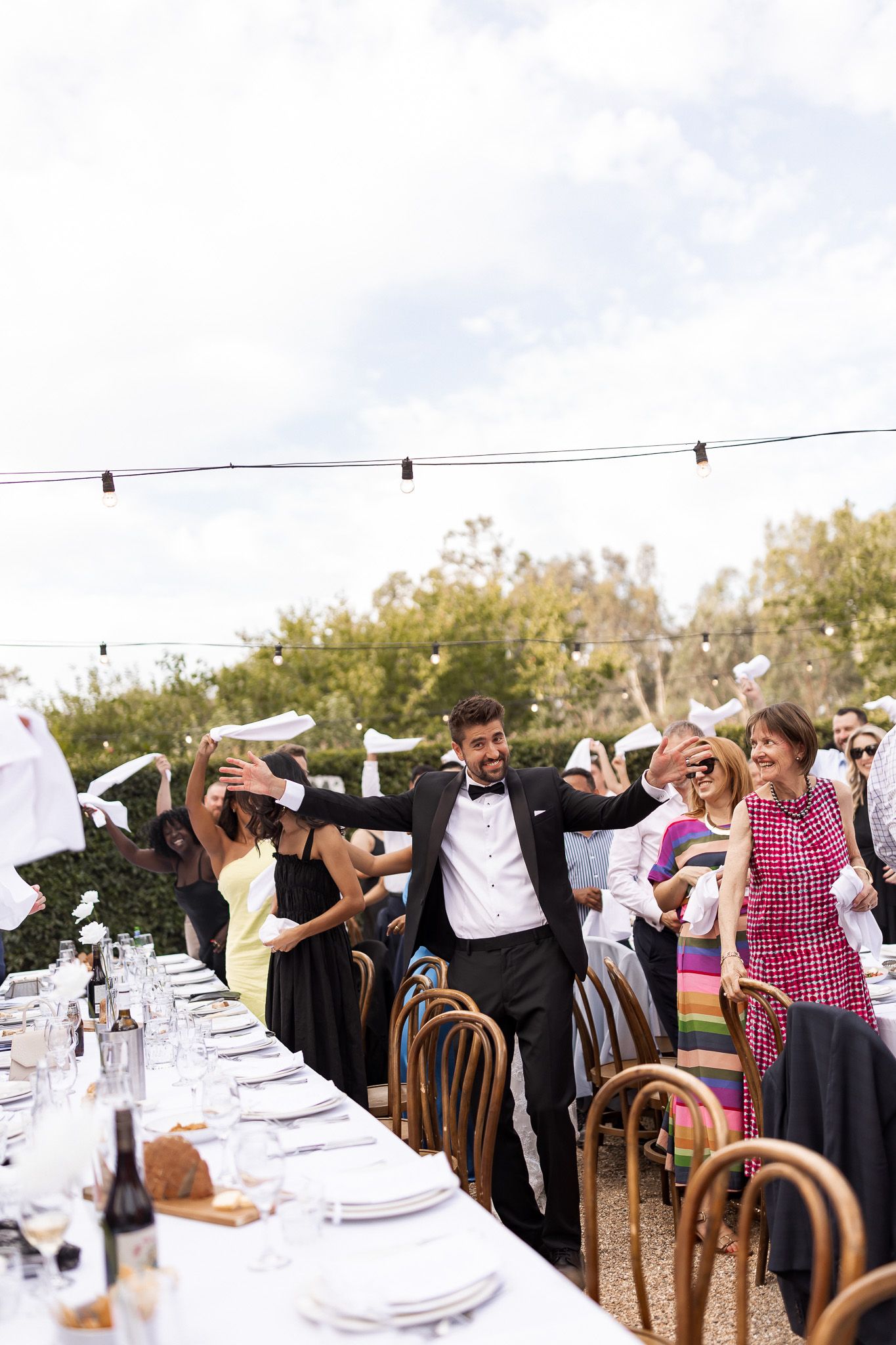 Groom in tuxedo celebrates with guests waving napkins at outdoor wedding reception dinner.