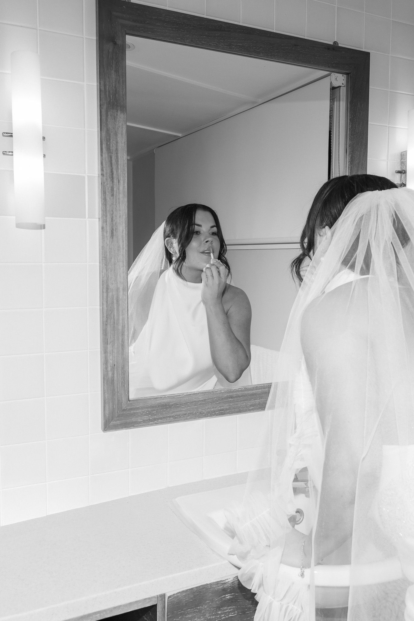Bride in wedding dress and veil applying makeup while looking into a wooden-framed bathroom mirror, black and white photo.