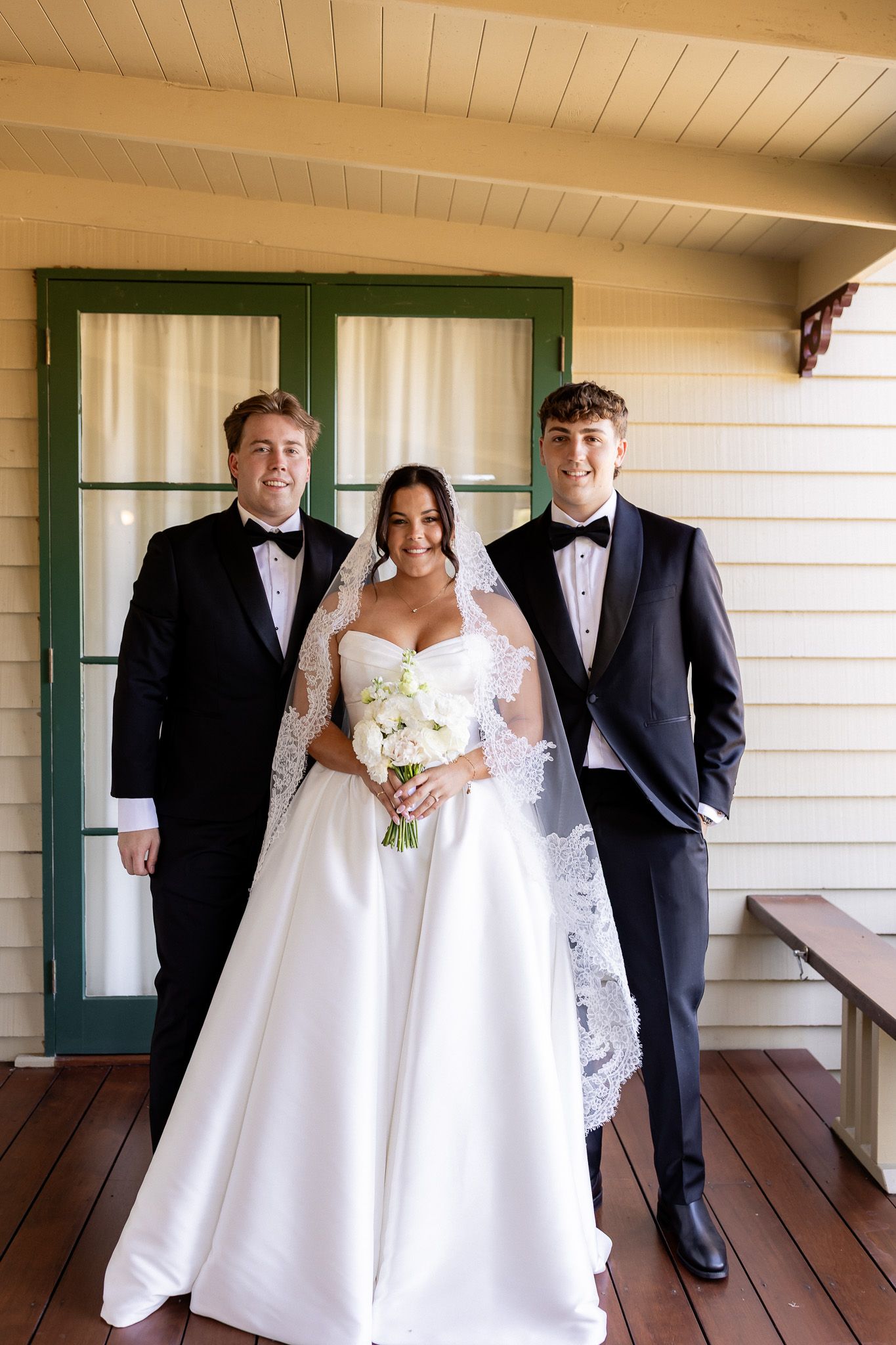 Bride in white gown with veil holding bouquet poses with two groomsmen in black tuxedos on a wooden porch.