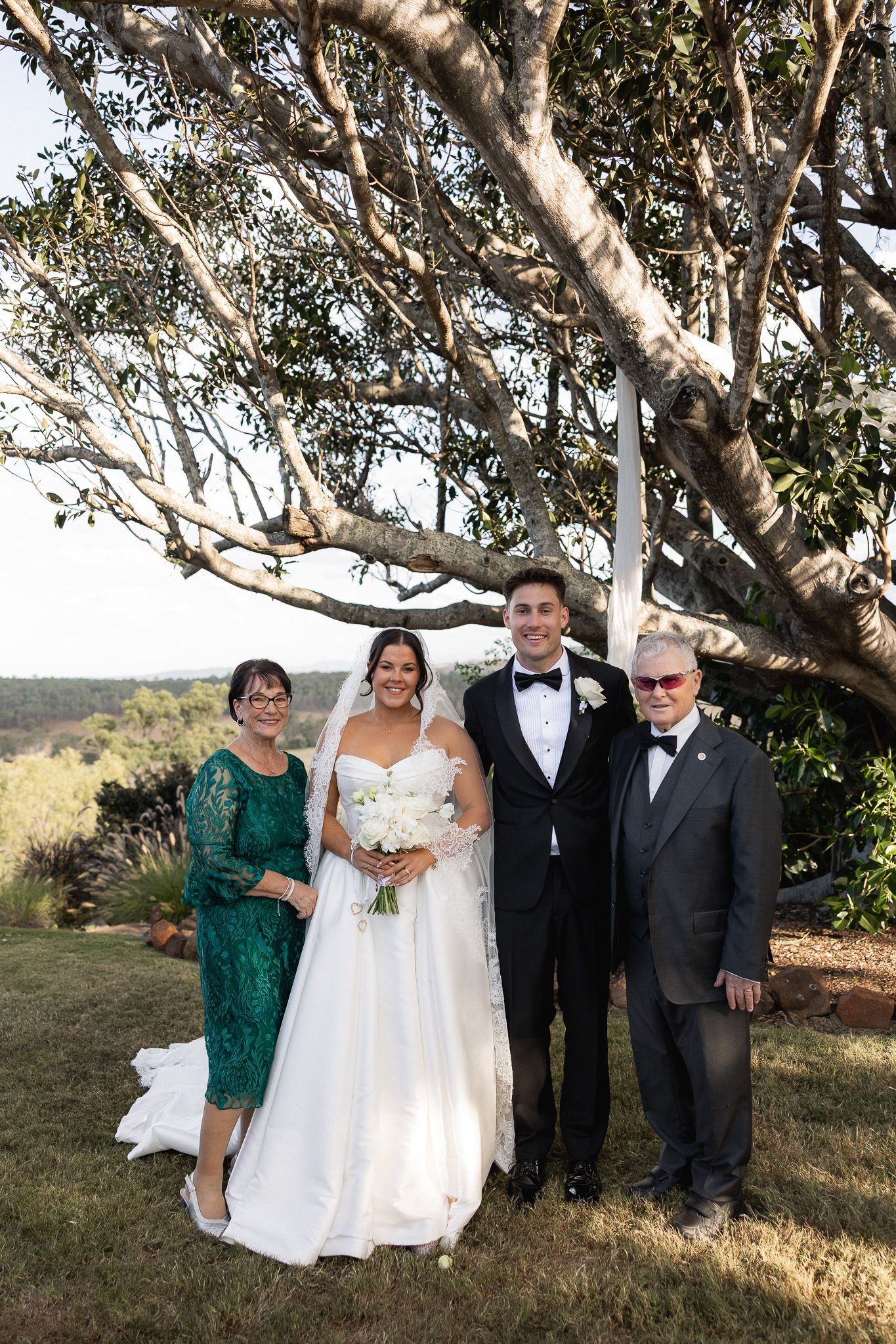 Bride and groom pose with two family members under a large tree in an outdoor garden setting.