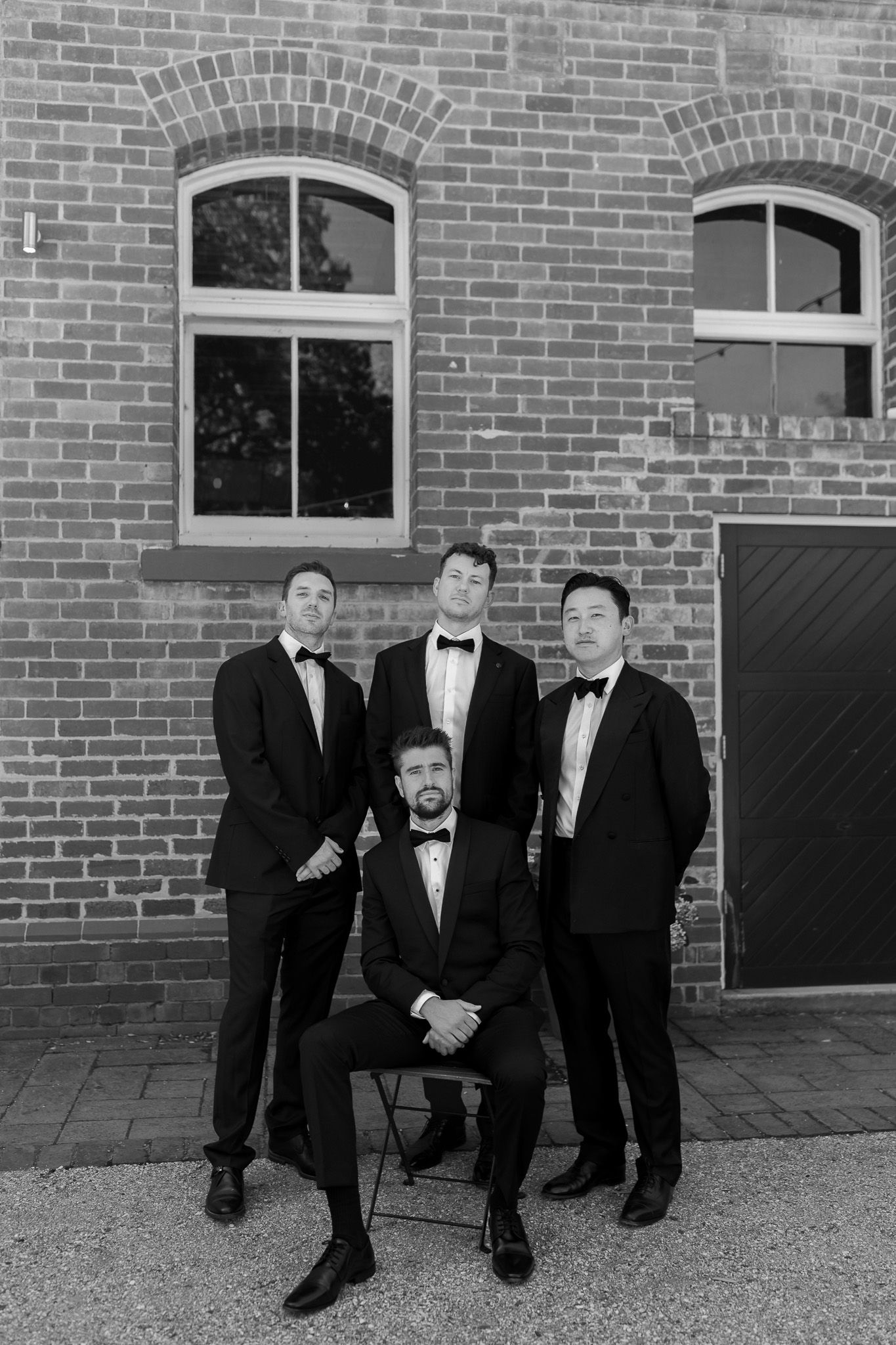 Four men in black tuxedos pose formally against a brick wall, one seated, in a black and white wedding photo.