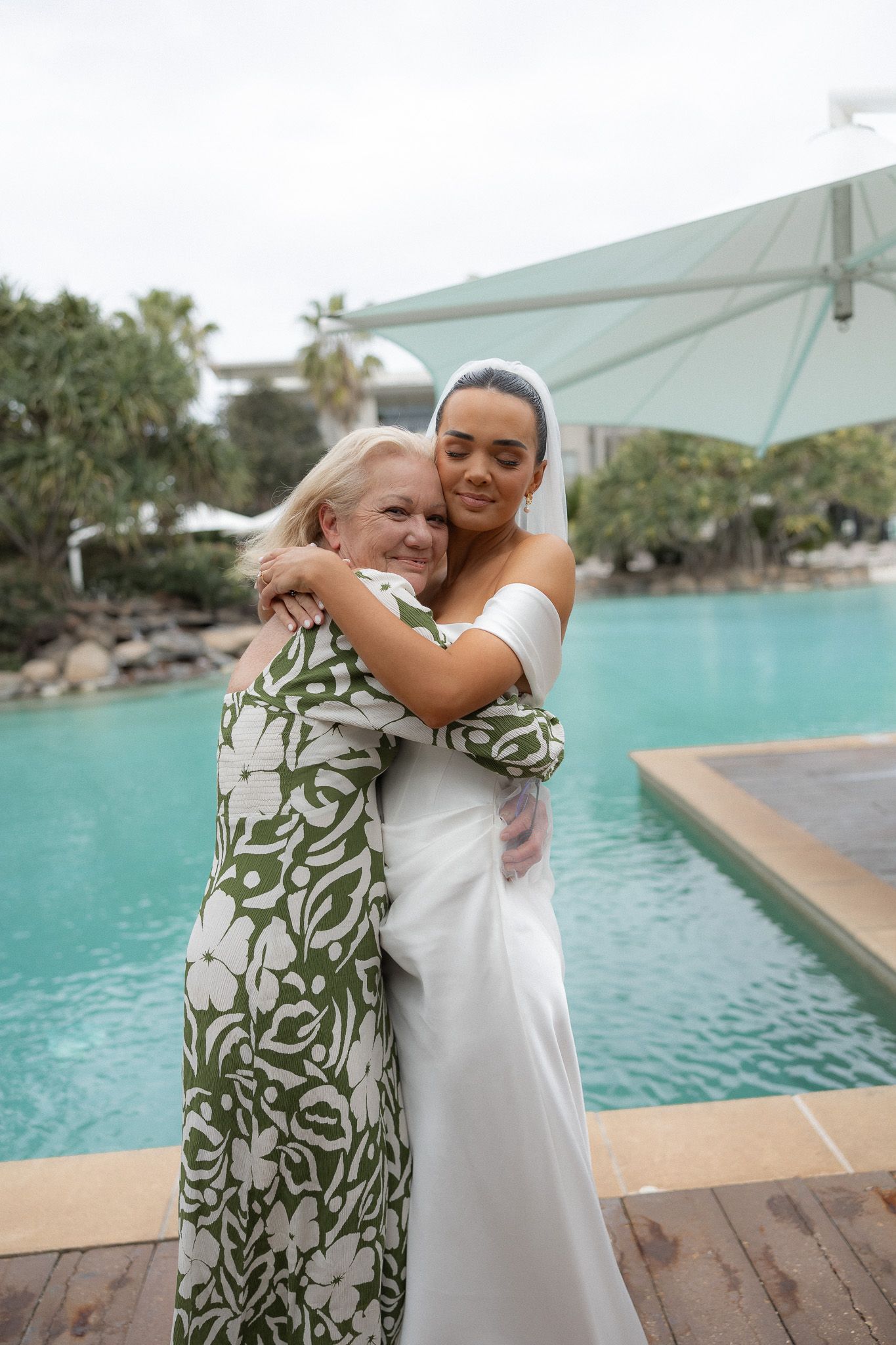 Bride in white gown hugs older woman in green floral dress poolside at outdoor wedding venue.