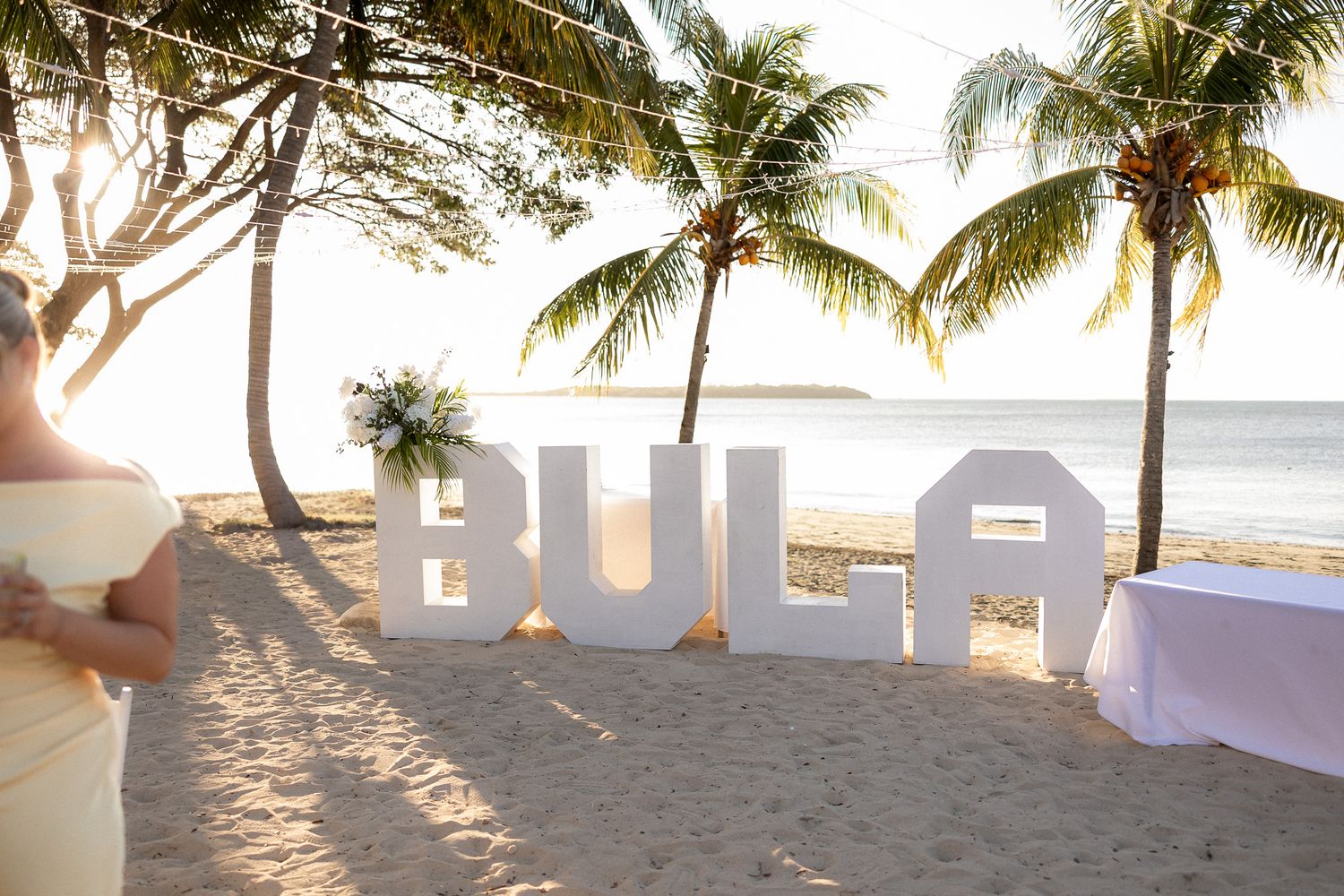 Large white BULA letters on sandy beach at sunset with palm trees and ocean backdrop.