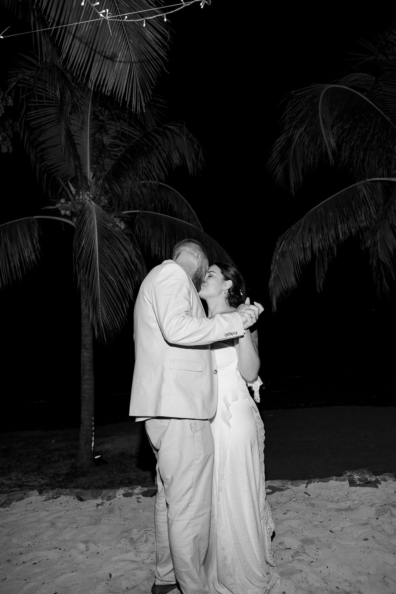 Black and white photo of bride and groom sharing a romantic kiss on the beach under palm trees at night.