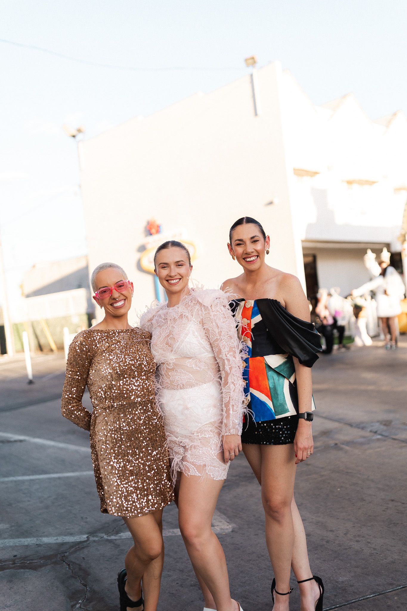 Three women in glamorous sequin and feathered dresses pose together at a Las Vegas event.