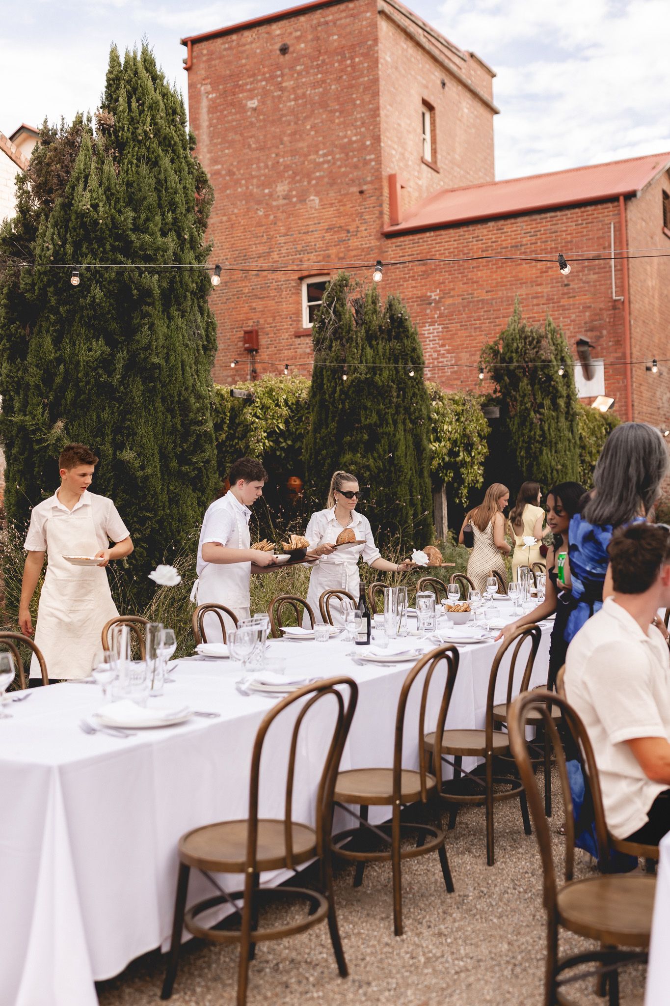 Catering staff prepare a long outdoor reception table at a rustic brick venue wedding.