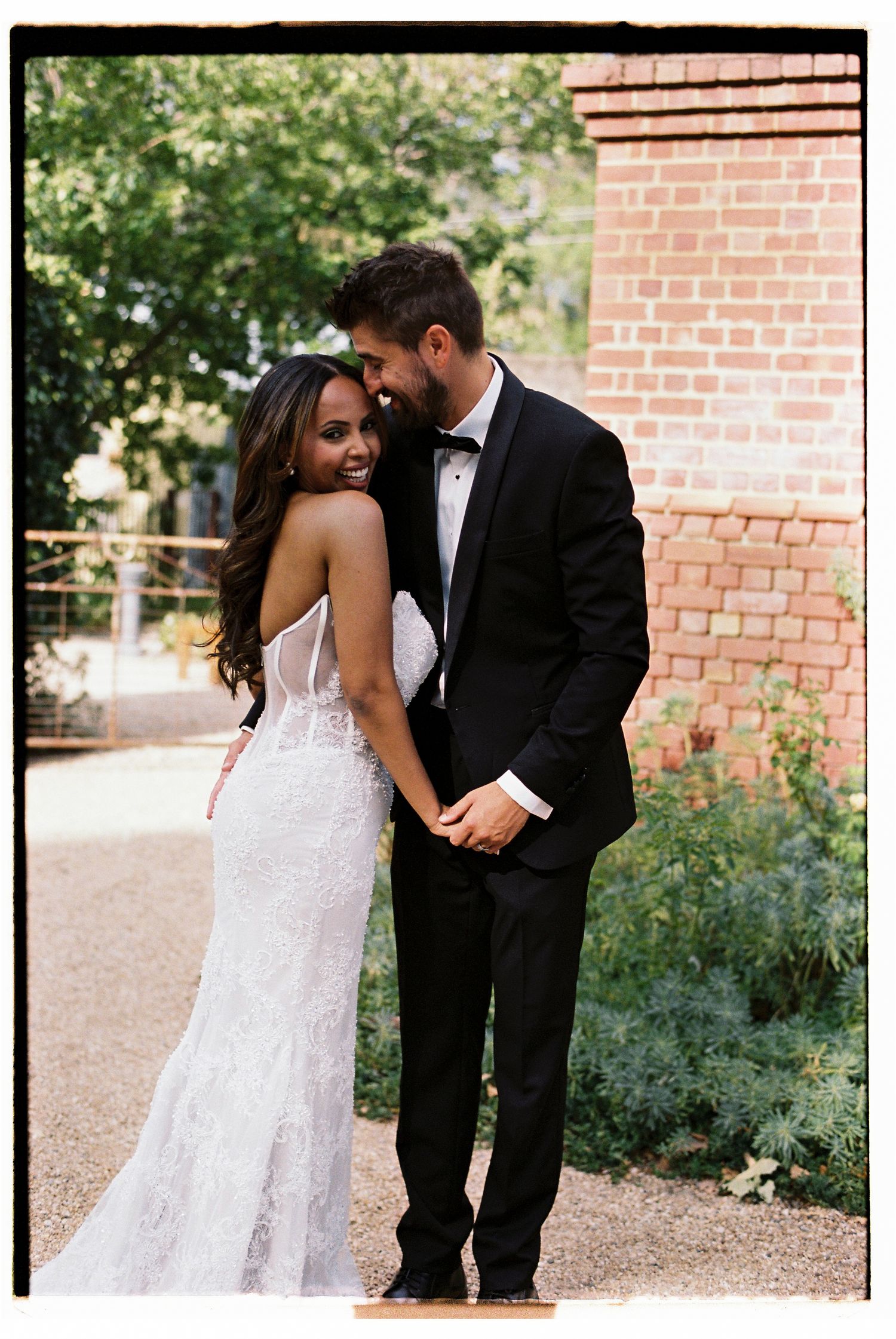 Bride in lace gown and groom in black tuxedo holding hands and smiling at outdoor wedding venue.