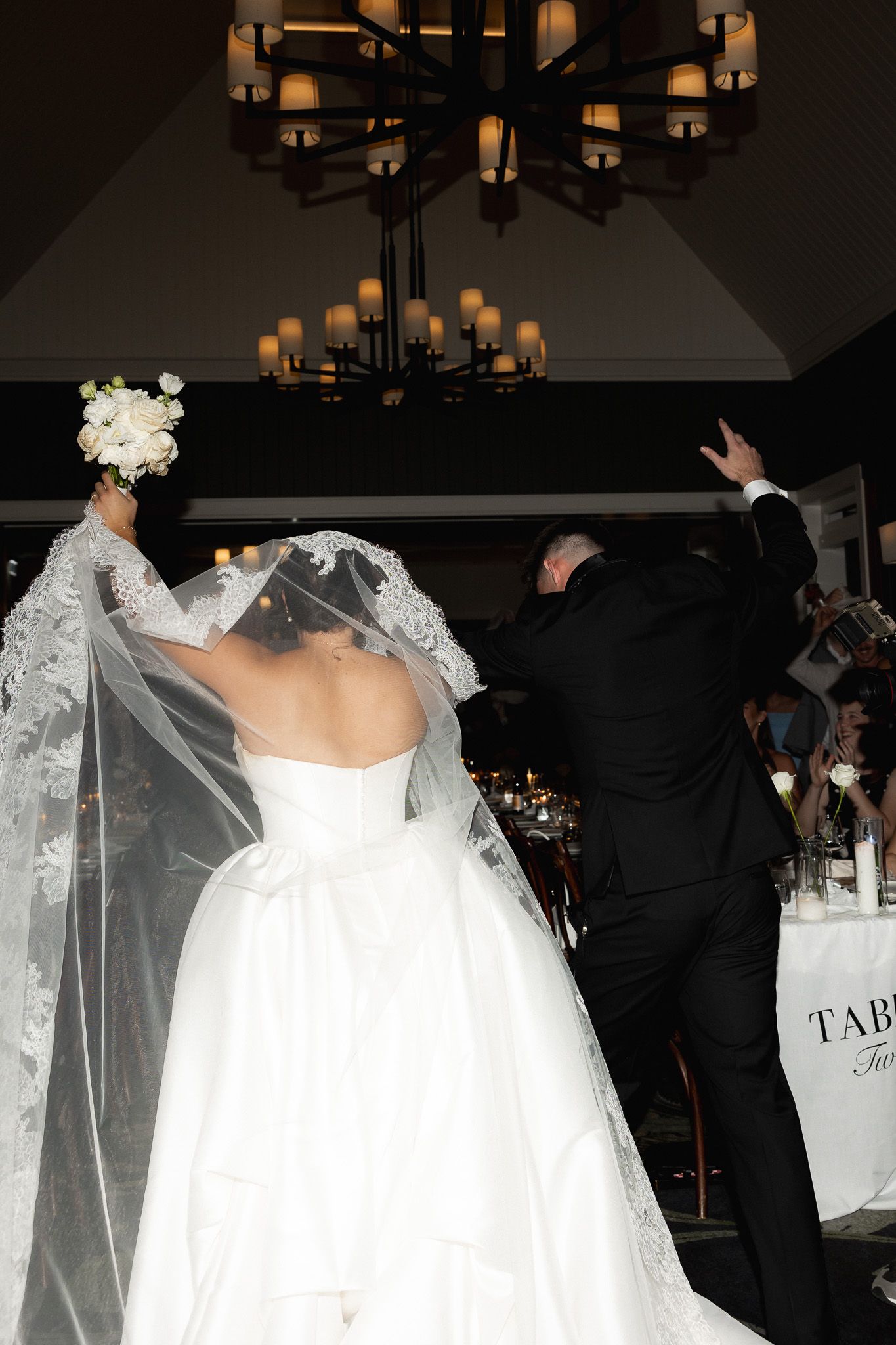 Bride in white ball gown raises bouquet triumphantly as couple makes grand entrance at wedding reception.