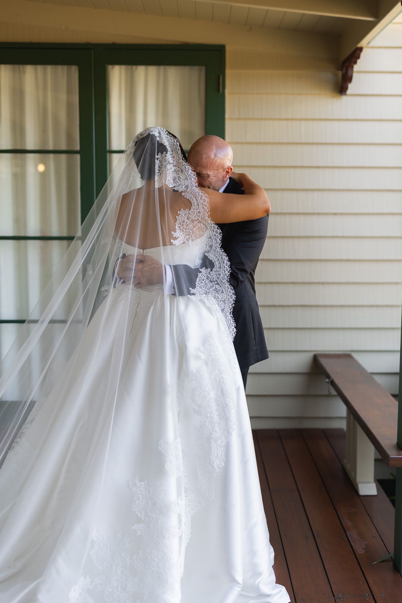 Bride in white lace gown and veil embracing a bald man on a wooden porch near green-trimmed windows.