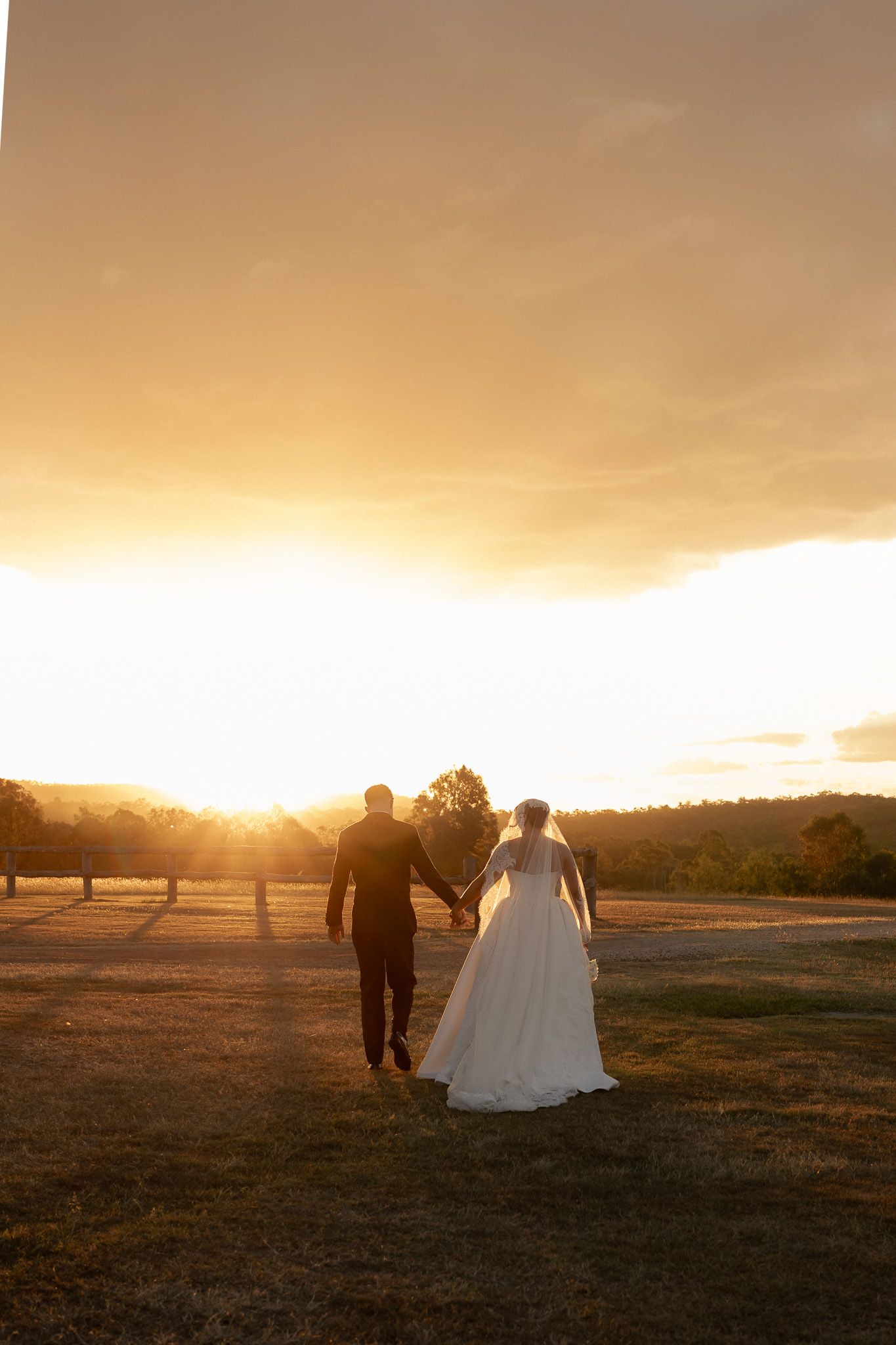 Bride and groom walk hand in hand toward a stunning golden sunset across an open field.
