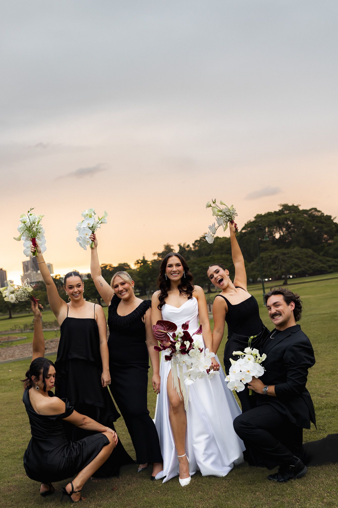 Bride in white gown poses with bridesmaids in black dresses holding bouquets at sunset outdoor wedding celebration.