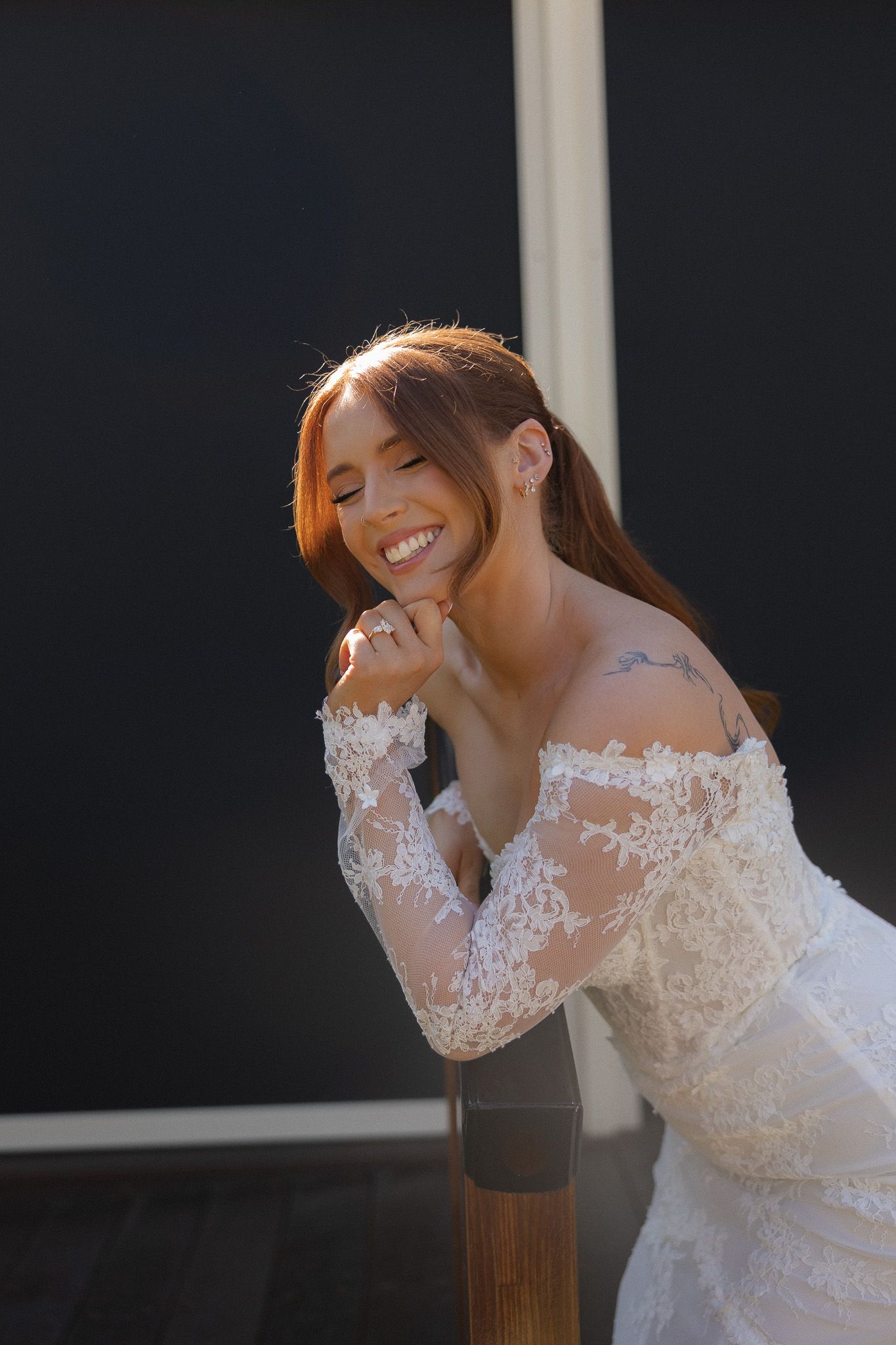Smiling bride in off-shoulder lace wedding dress with tattoo, leaning on railing in warm sunlight.