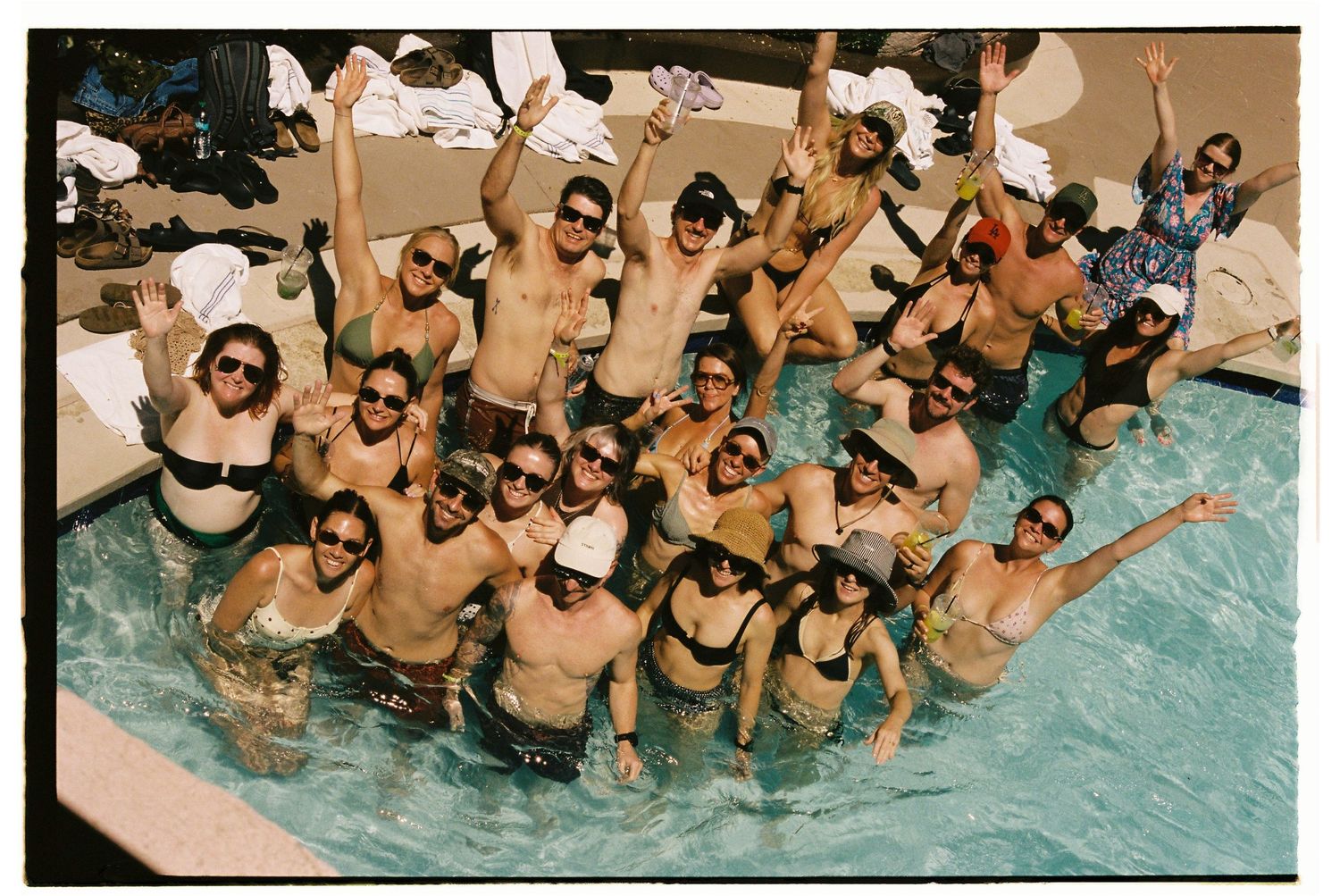 Large group of smiling people in swimwear posing together in an outdoor pool, raising arms and celebrating.