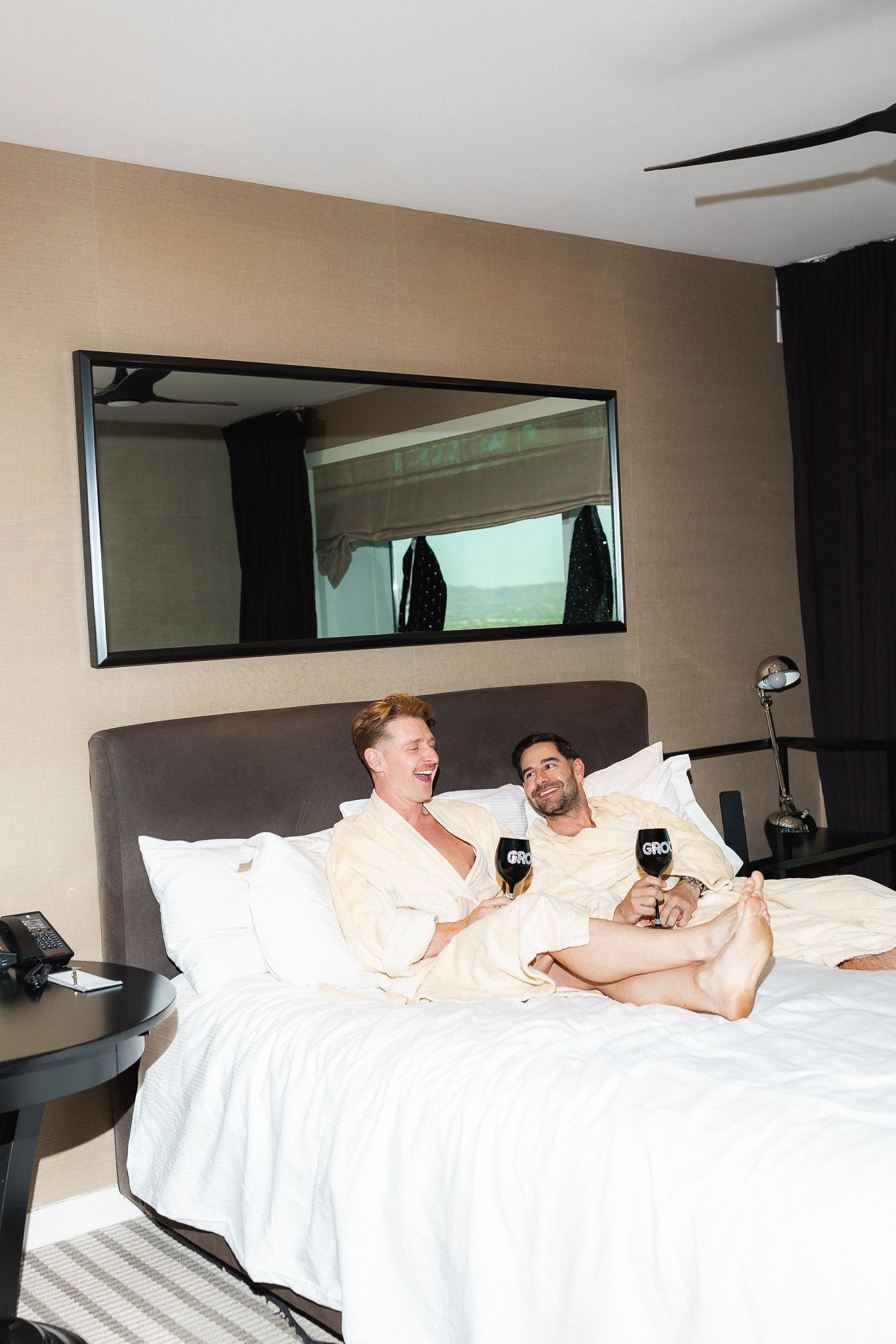 Two men in white robes relaxing on a hotel bed holding branded mugs, laughing together.