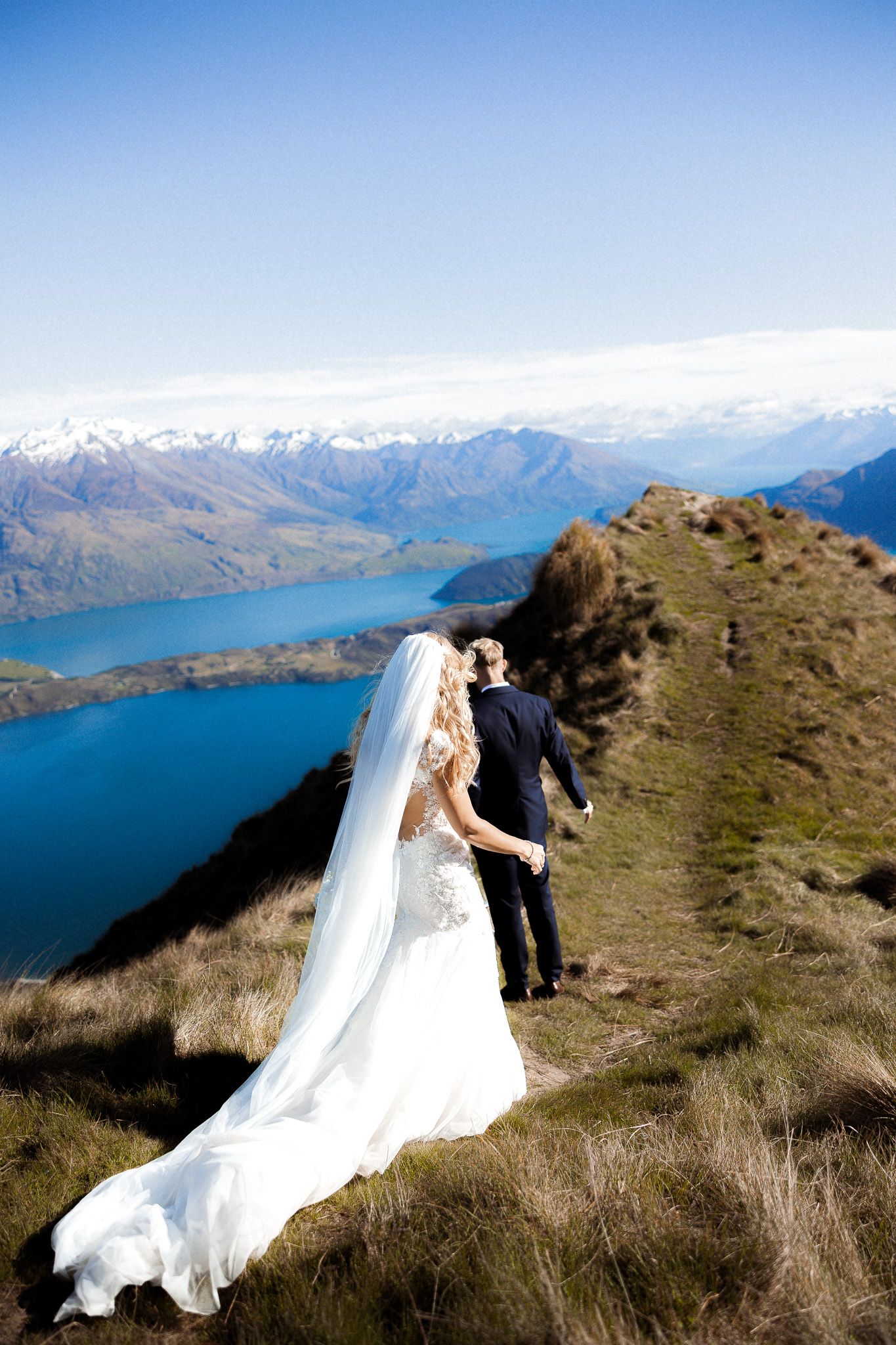 Bride and groom stand on a mountain ridge overlooking a stunning blue lake and snow-capped peaks on their wedding day.