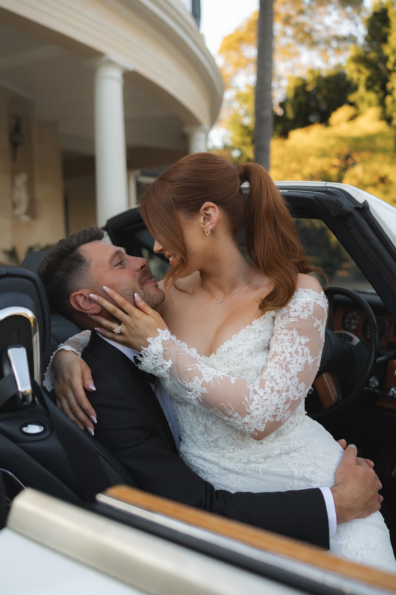 Bride in lace gown and groom in suit share a romantic moment in a white convertible car on their wedding day.