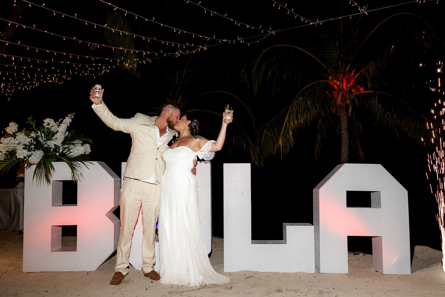 Couple celebrating wedding on beach at night beside illuminated BULA letters with string lights overhead.