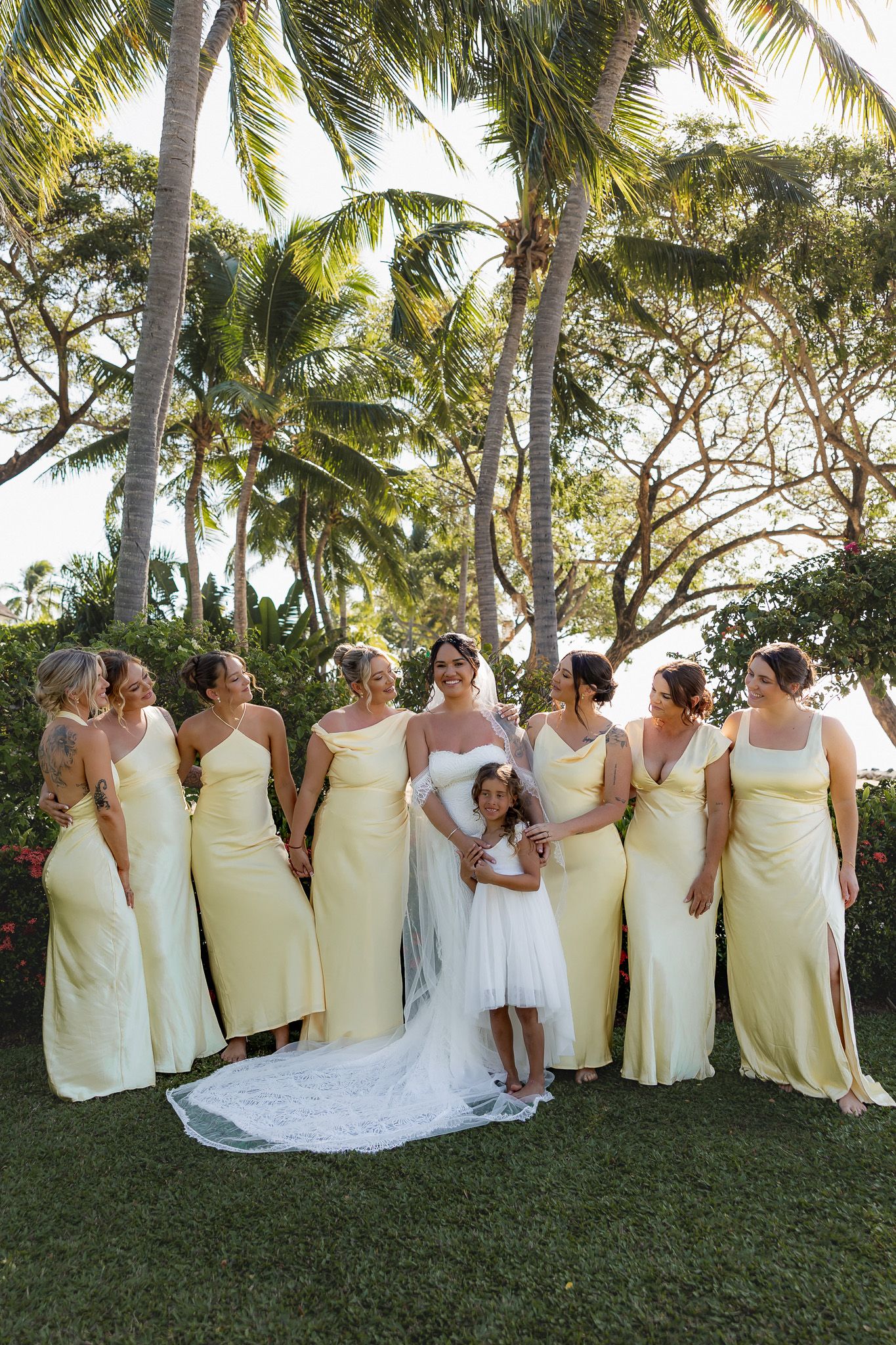 Bride with bridesmaids in yellow dresses and flower girl posing under tall palm trees on green lawn.