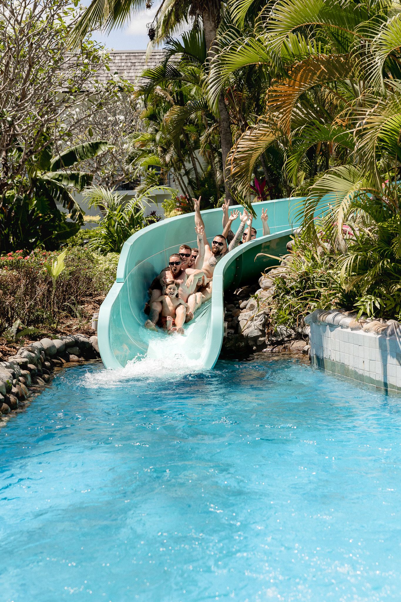 Group of people laughing and splashing down a tropical water slide into a bright blue pool.