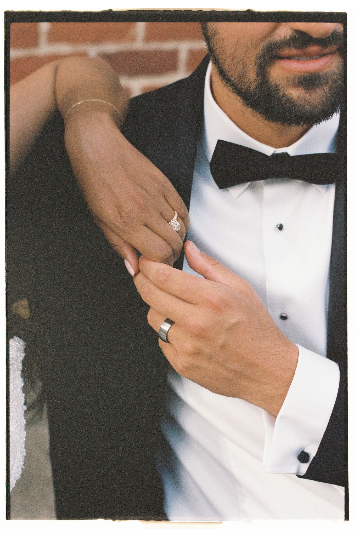 Bride and groom holding hands showing wedding rings, groom in black tuxedo with bow tie, film photography style.
