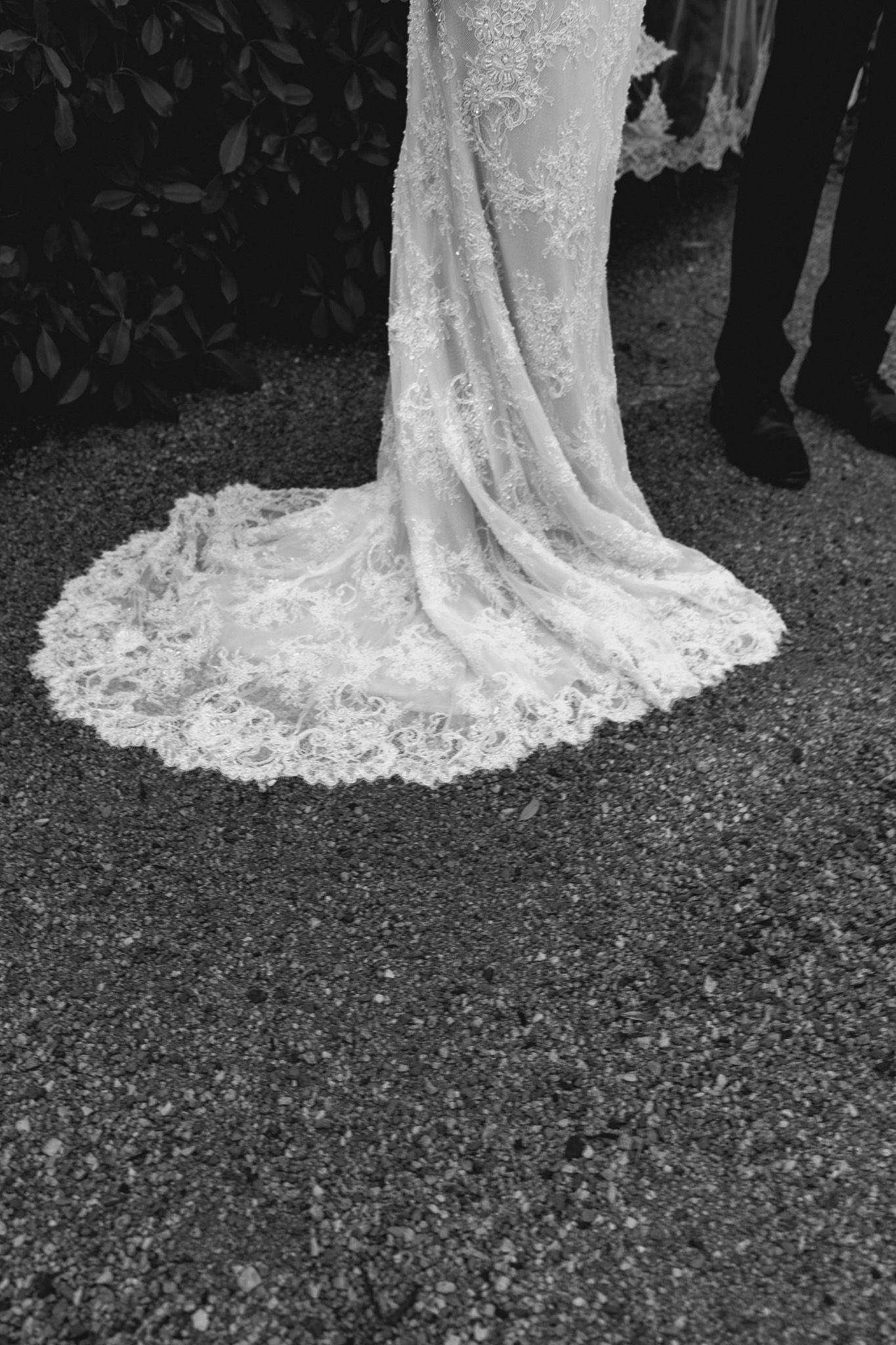 Close-up black and white photo of an intricate lace wedding dress train spread on gravel.
