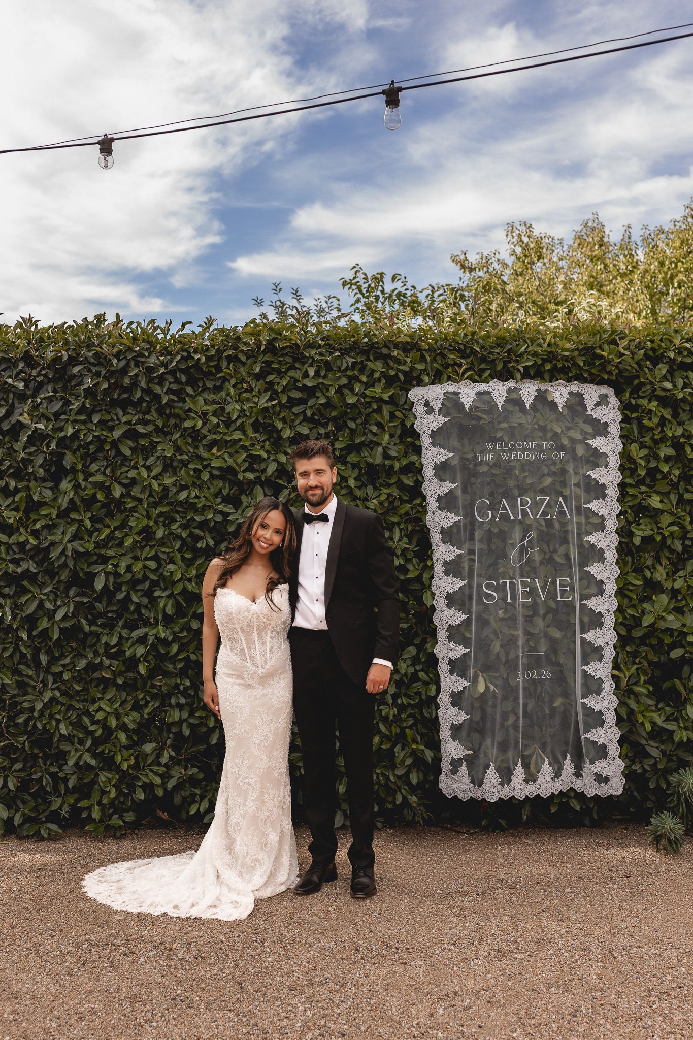 Bride in lace gown and groom in tuxedo pose beside wedding welcome sign reading Garza and Steve against green hedge backdrop.