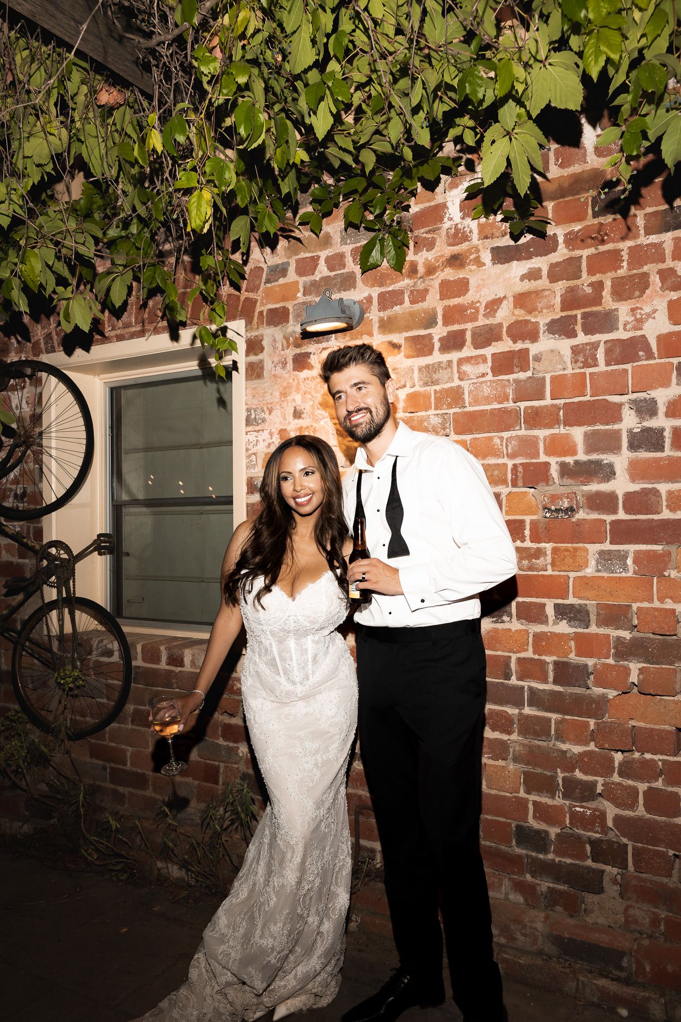 Smiling bride and groom posing together against a rustic brick wall during their nighttime wedding reception.