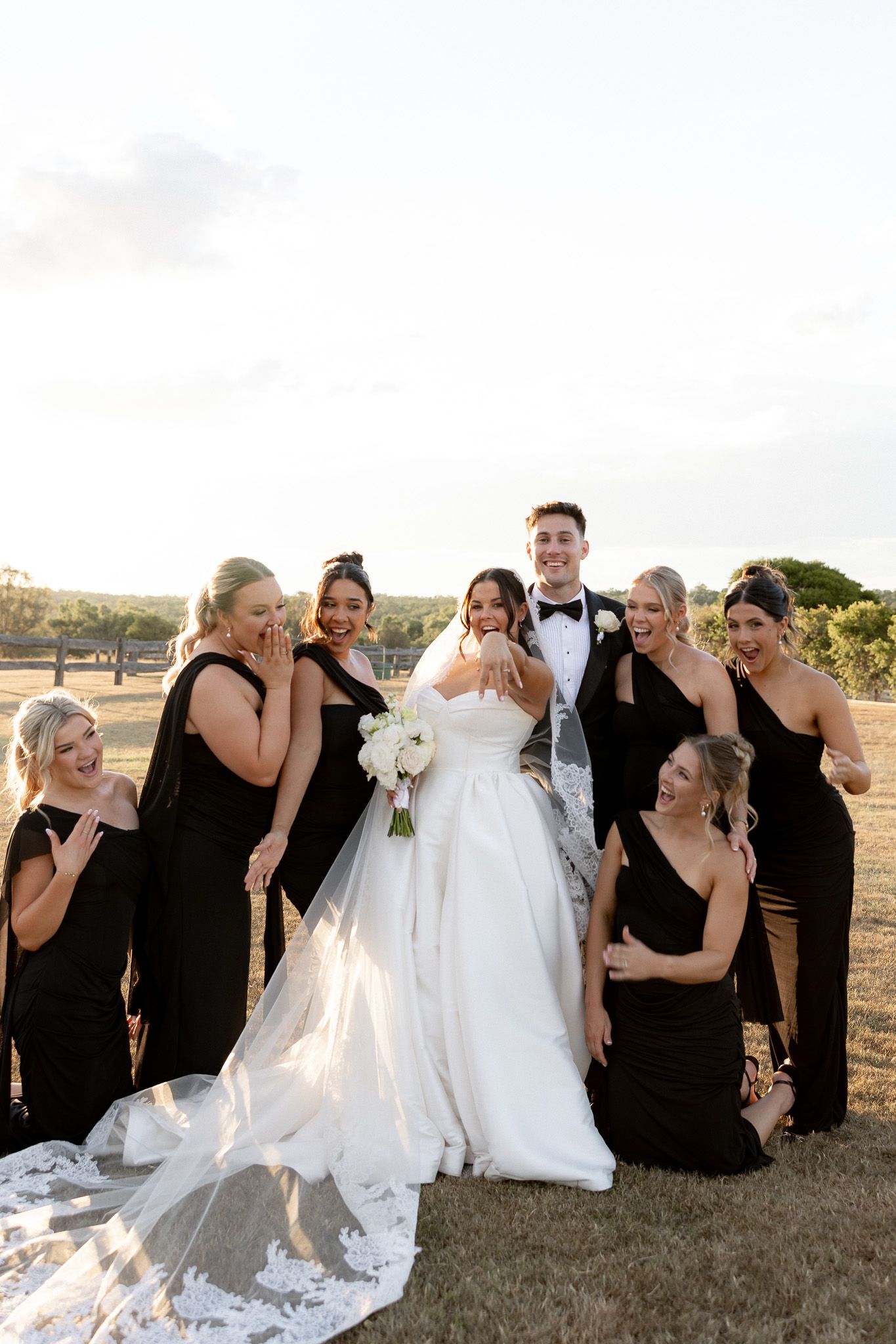 Bride and groom surrounded by bridesmaids in black dresses laughing outdoors at golden hour on wedding day.