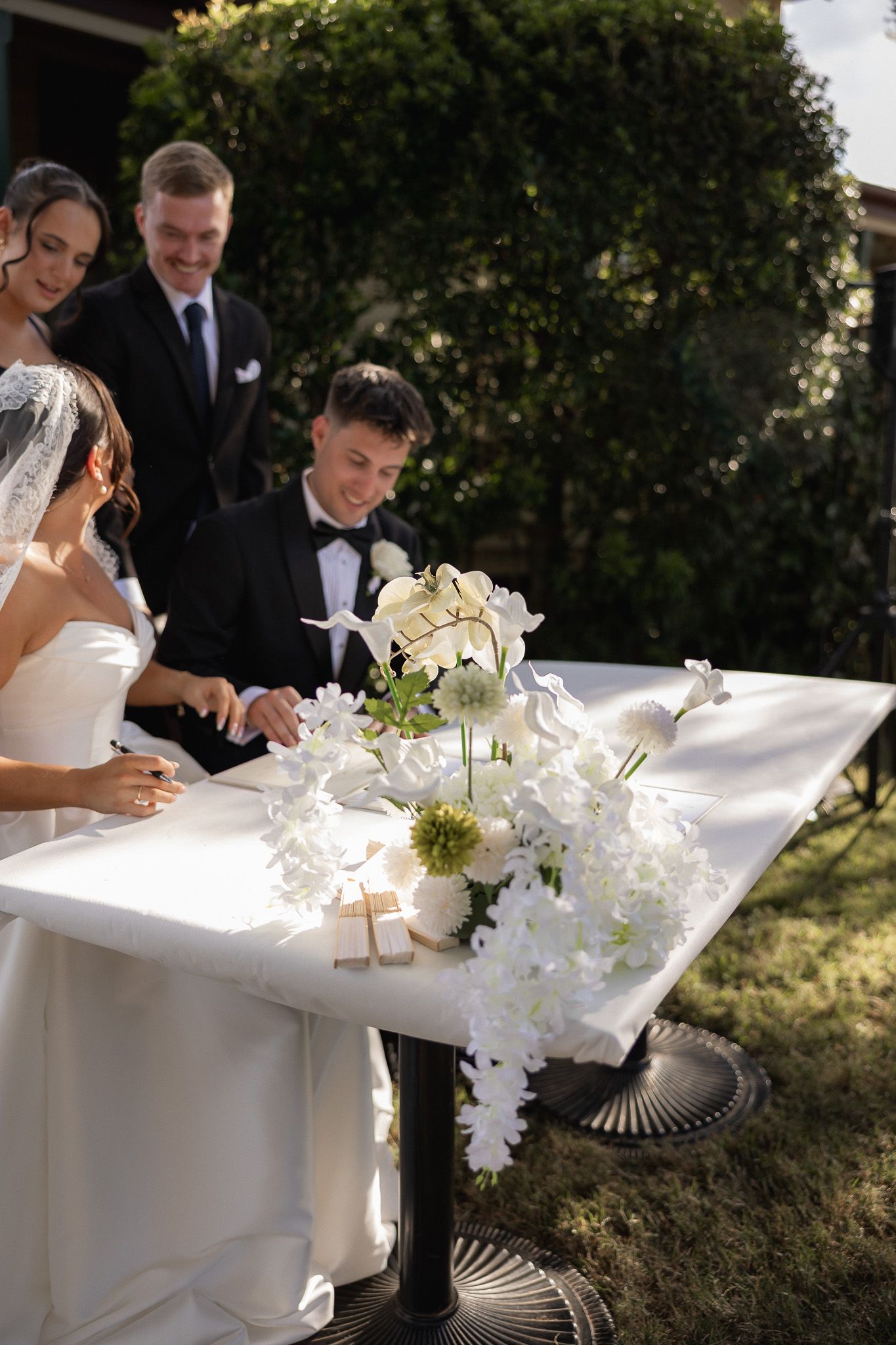 Bride and groom sign wedding certificate at floral decorated table with witnesses smiling.
