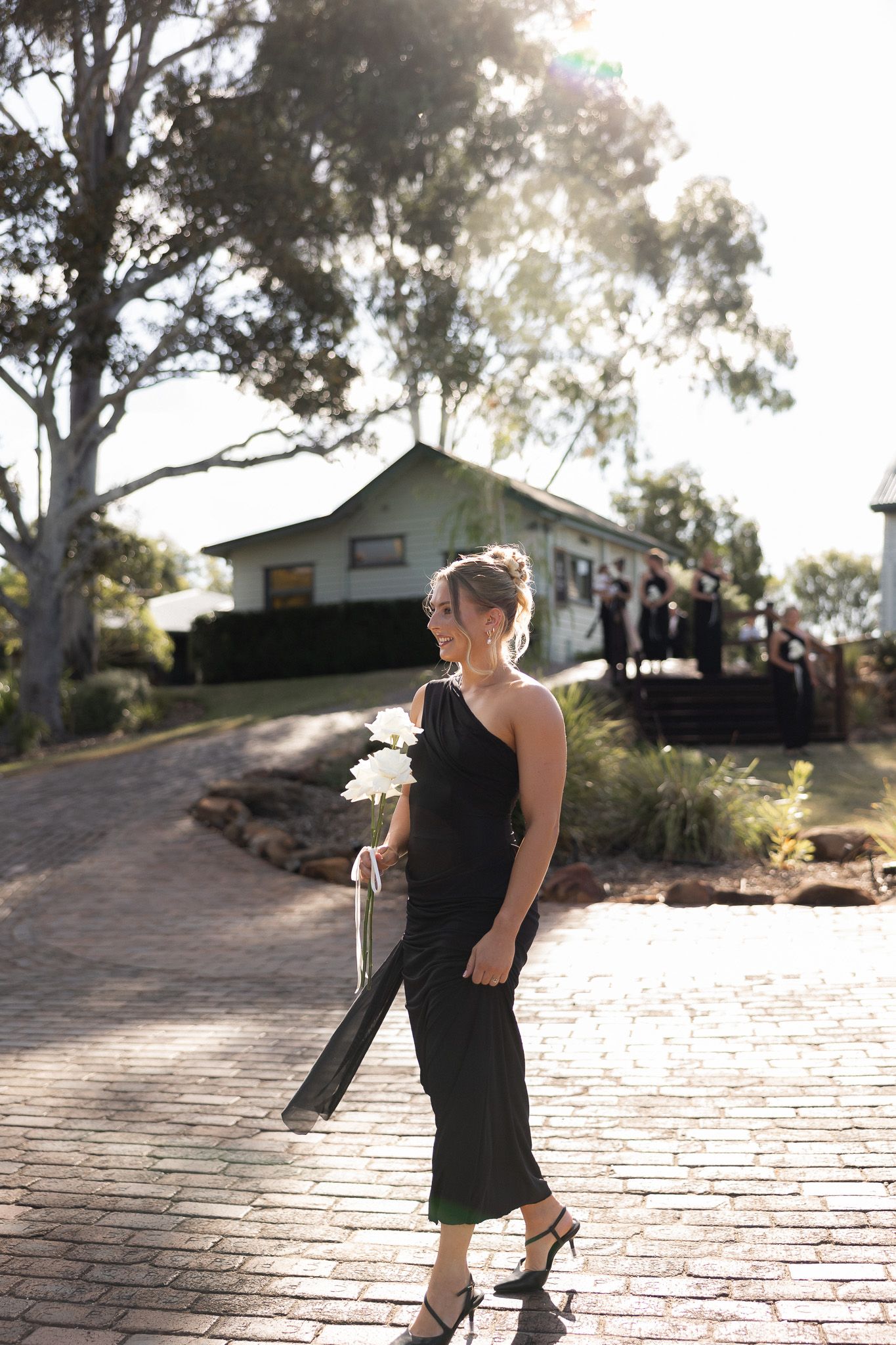 A bridesmaid in a black one-shoulder dress walks along a brick path holding white flowers at an outdoor wedding venue.