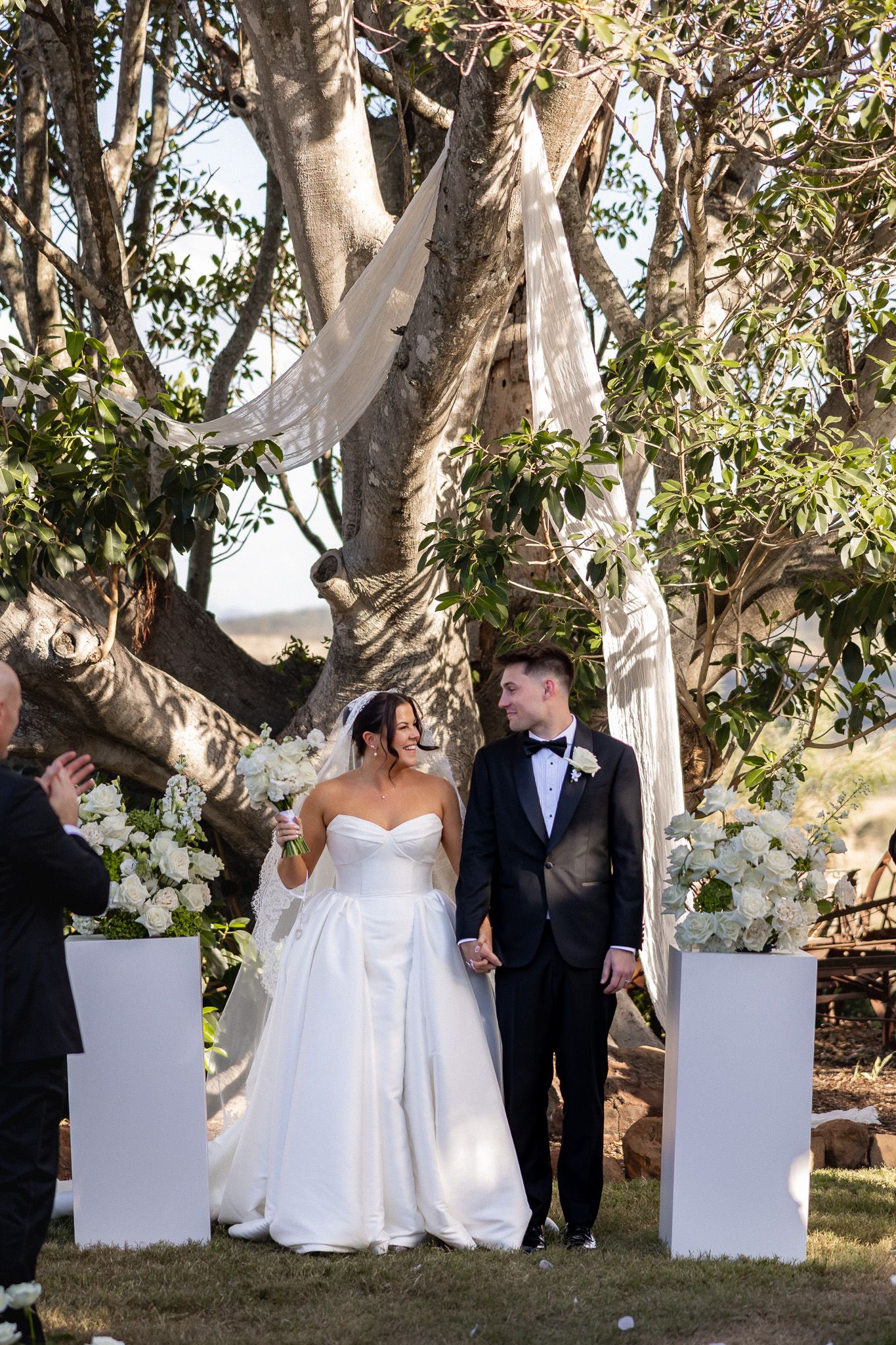 Bride and groom holding hands at outdoor wedding ceremony with white floral arrangements and draped fabric on trees.