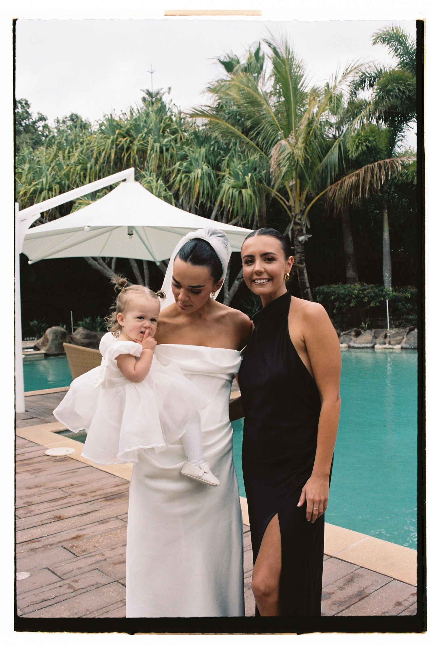 Bride holding baby in white dress poses with guest in black gown by tropical pool.