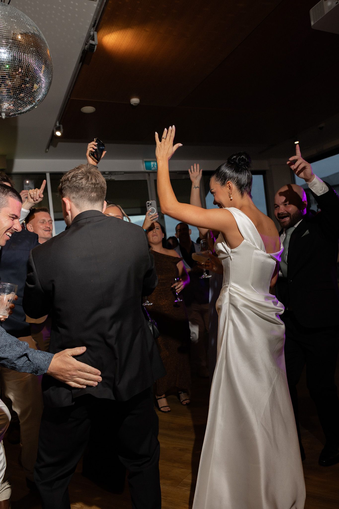 Bride in white satin gown celebrates with hands raised on crowded dance floor at wedding reception party.