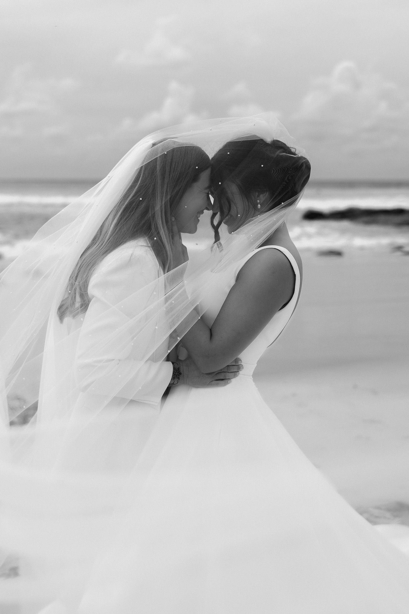 Black and white romantic photo of two brides embracing under a veil on beach.