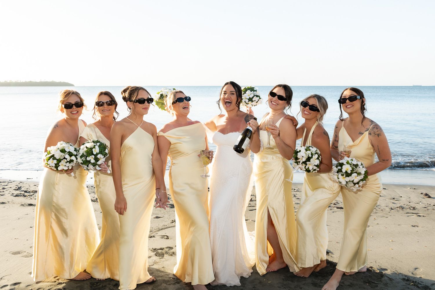 Bride and bridesmaids in yellow dresses wearing sunglasses laughing on sandy beach with calm ocean behind.