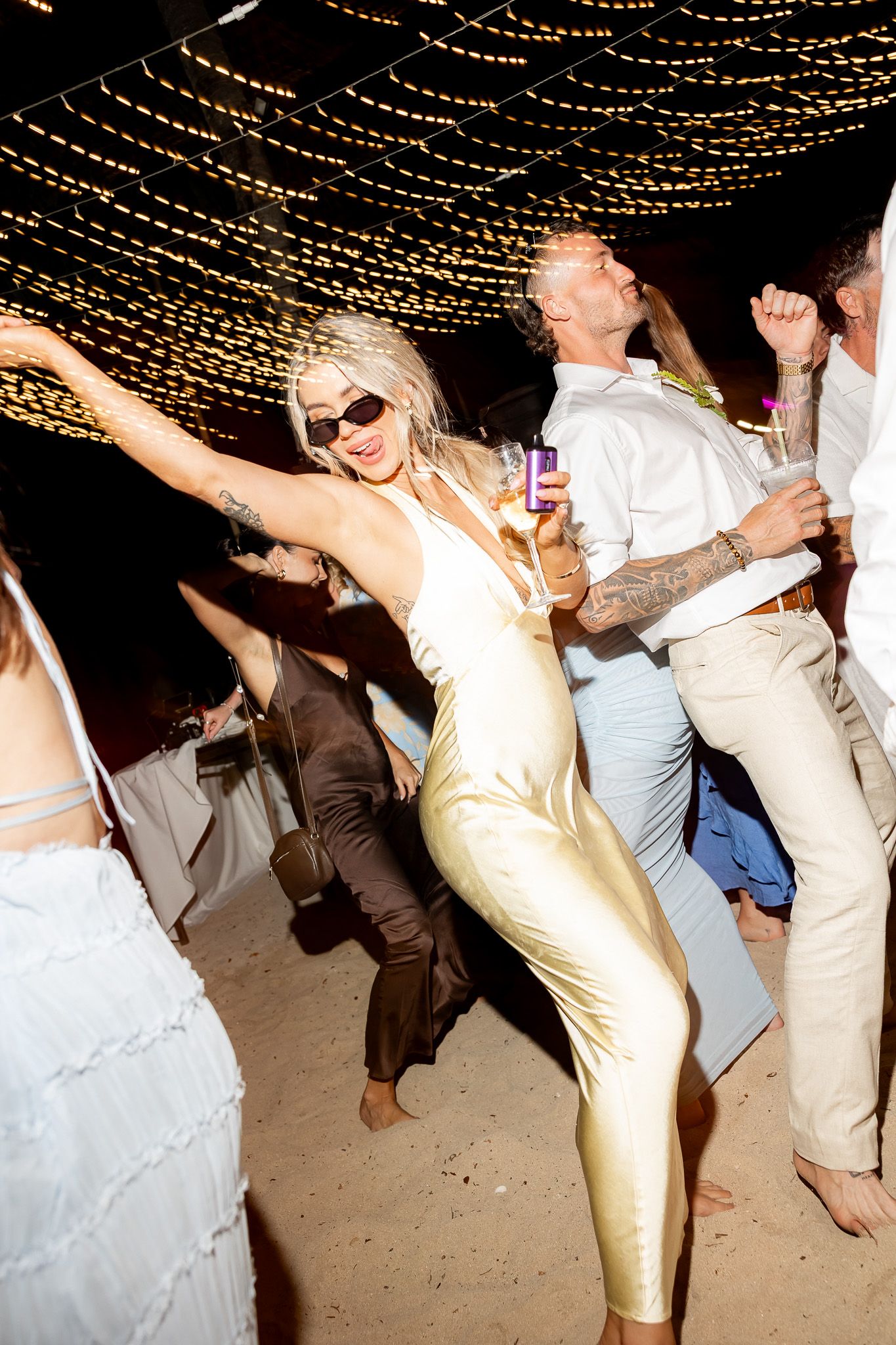 Guest in a gold satin dress dances energetically under string lights at an outdoor beach wedding reception.