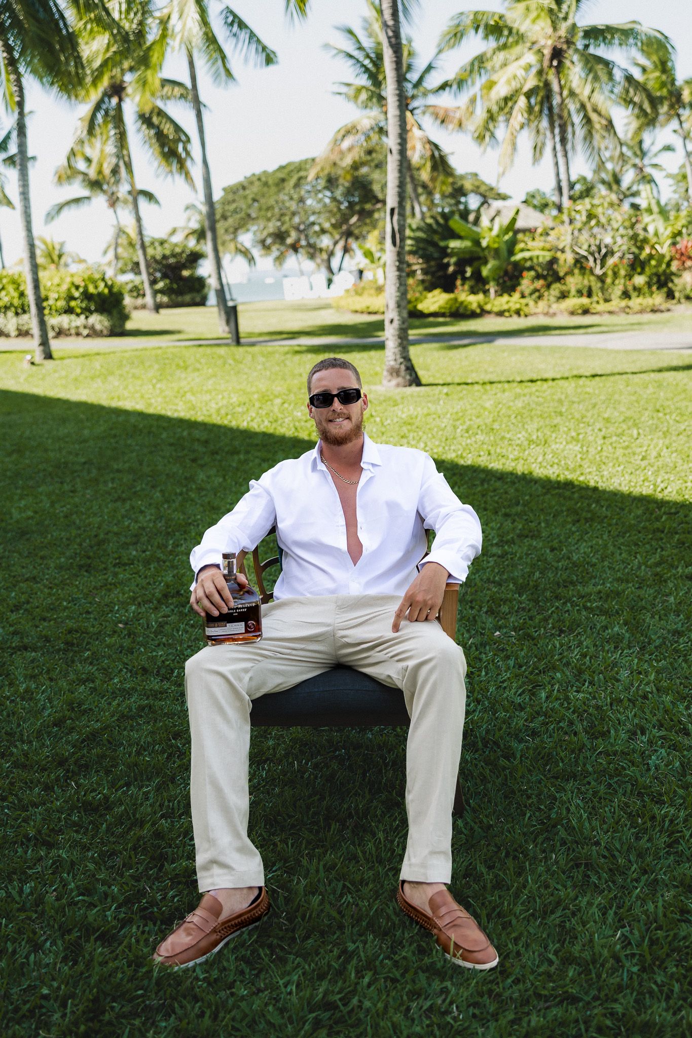 Stylish man in white shirt and khaki pants sitting on chair holding whiskey bottle on tropical lawn.
