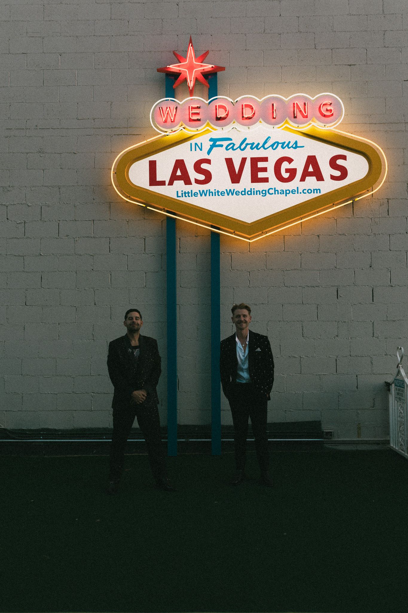 Two grooms stand beneath a glowing Wedding in Fabulous Las Vegas neon sign at Little White Wedding Chapel.