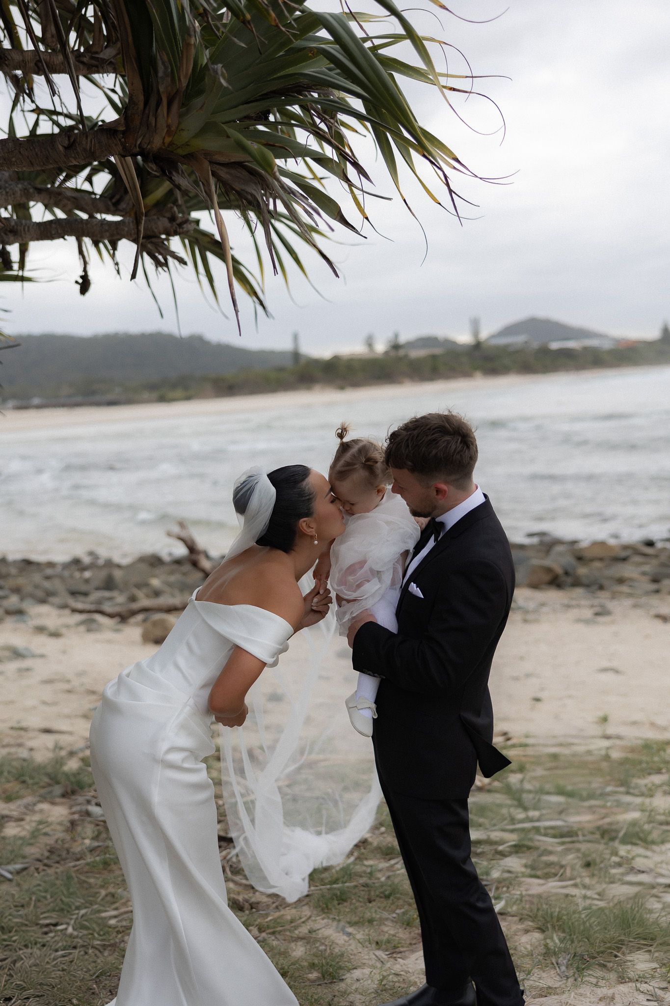 Bride and groom kissing their baby on a beach wedding day with palm trees and ocean in background.