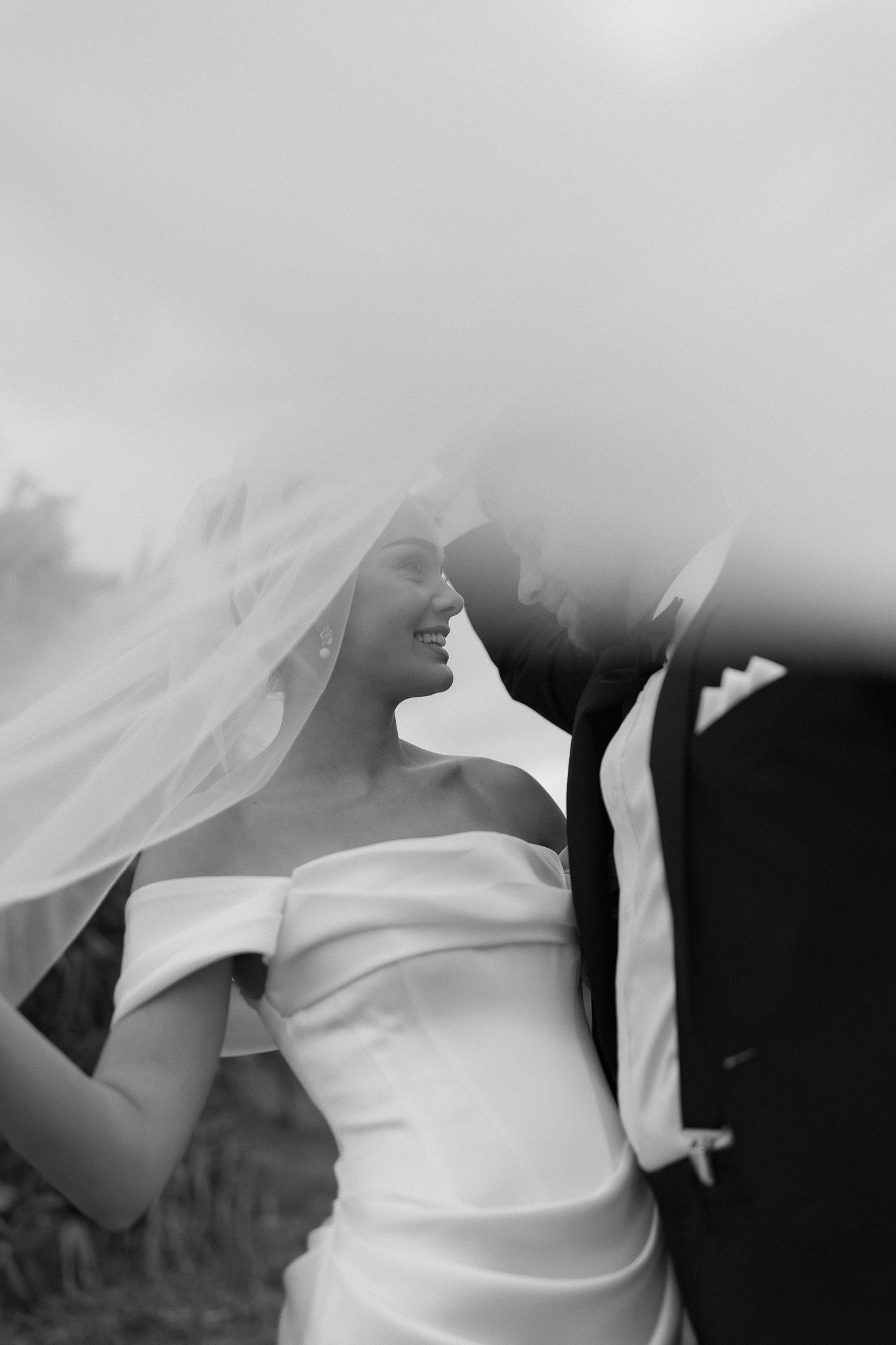 Black and white romantic portrait of smiling bride in off-shoulder gown under flowing veil.