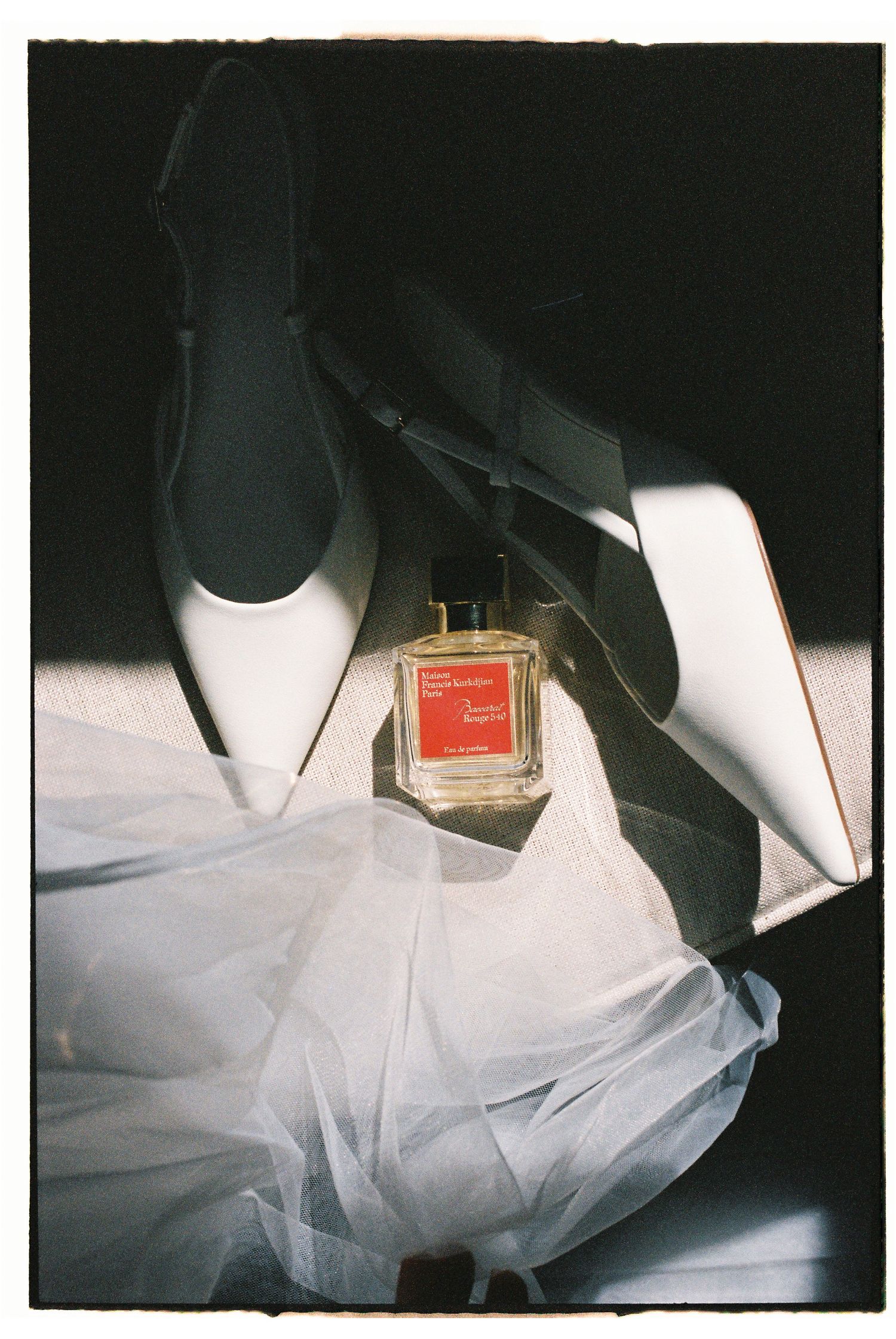 Maison Francis Kurkdjian perfume bottle resting on white bridal heels and veil in dramatic light.