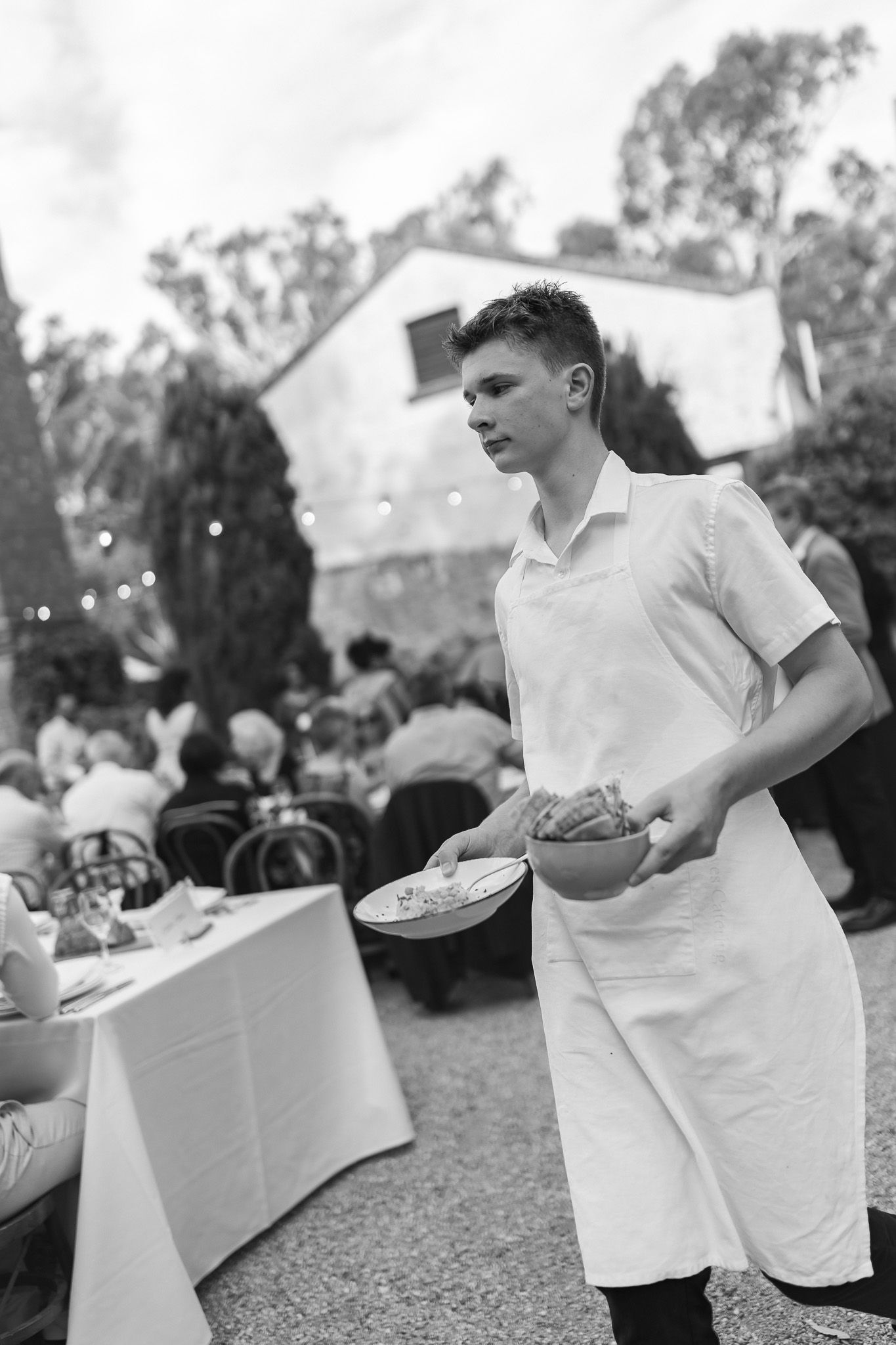 Black and white photo of a young waiter in white apron serving food at an outdoor wedding reception.