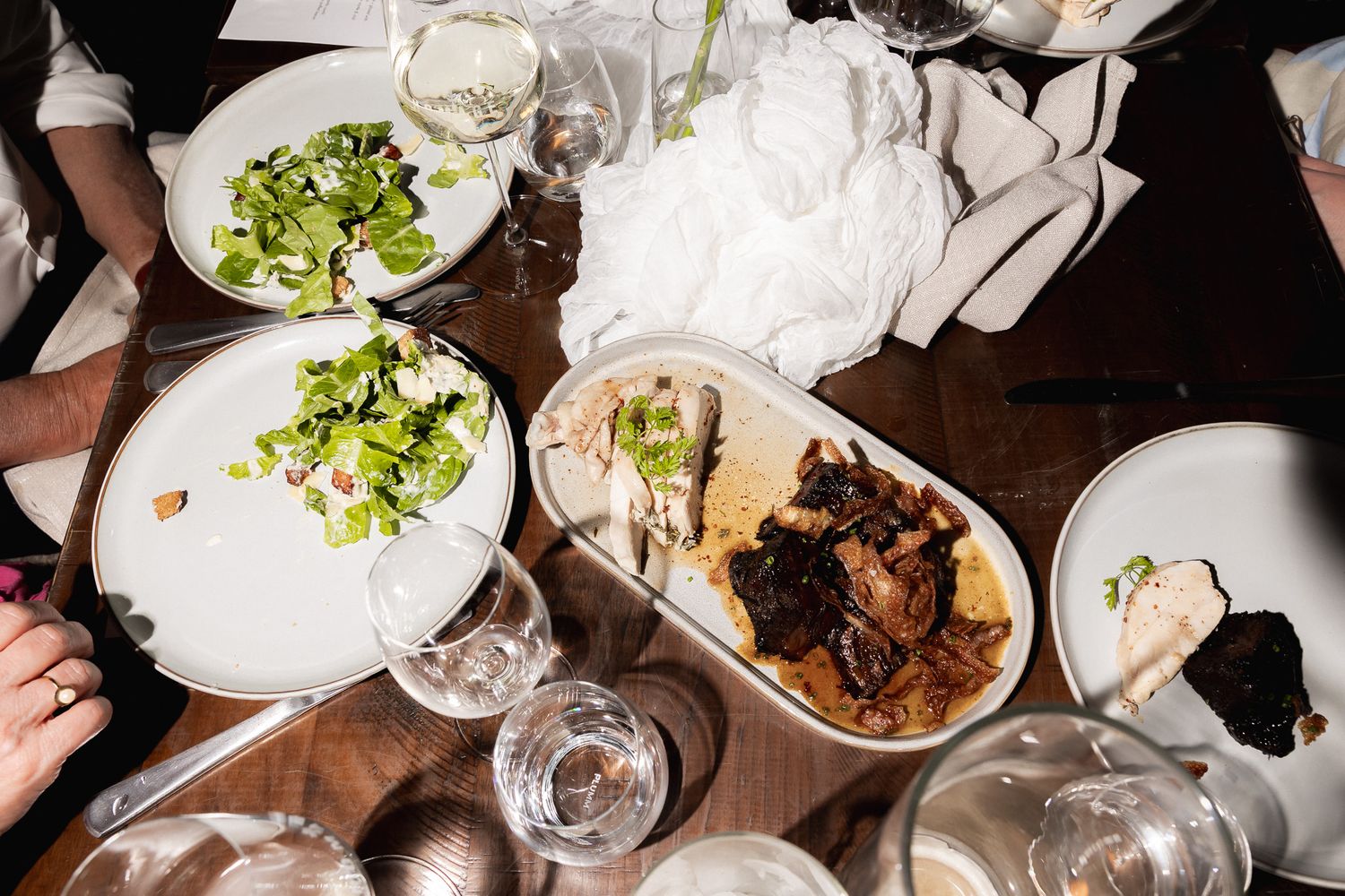 Overhead view of wedding reception dinner table with salad, roasted meat and empty wine glasses.