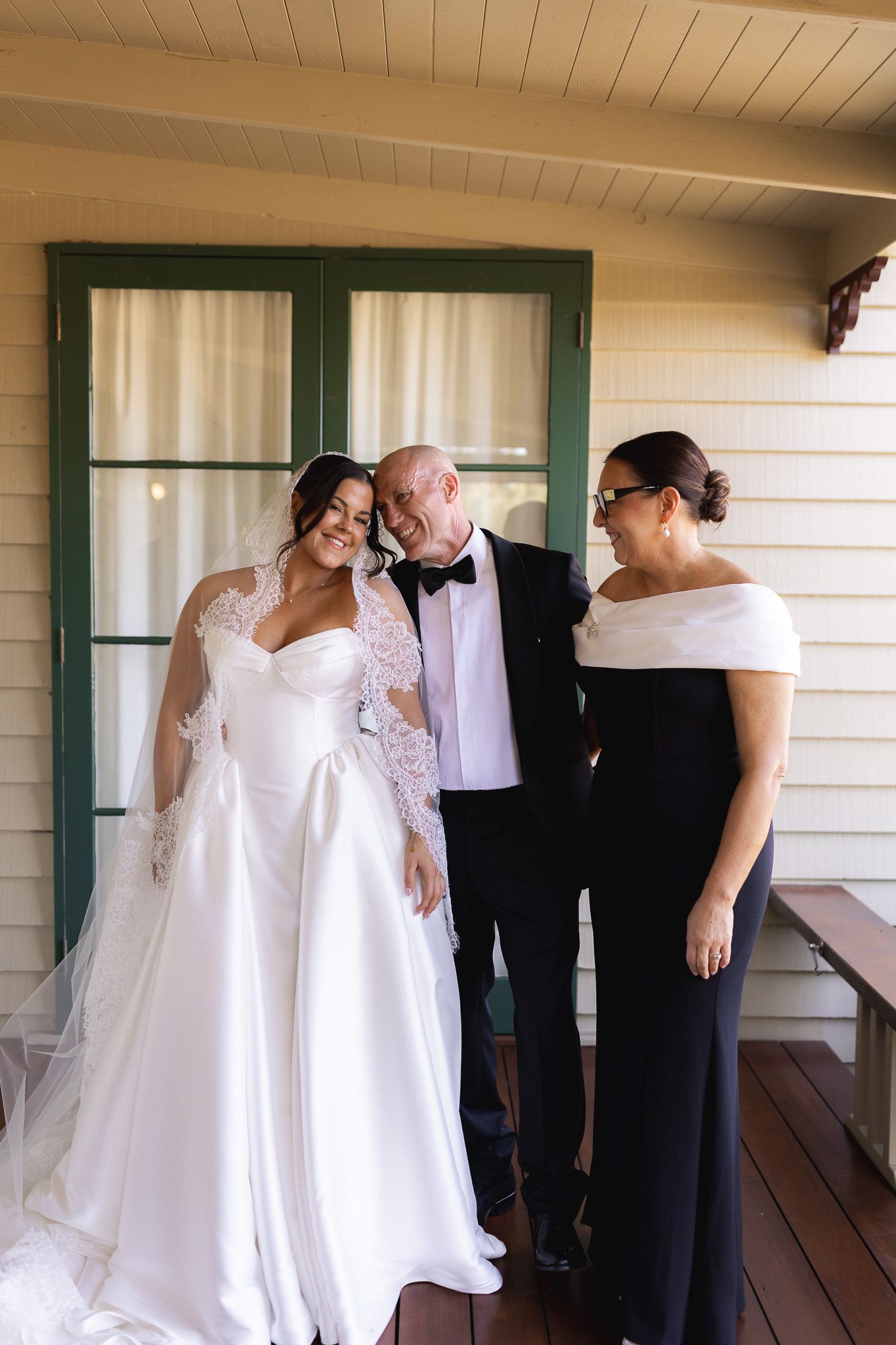 Bride in white lace gown poses with older man in tuxedo and woman in black dress on wooden porch.