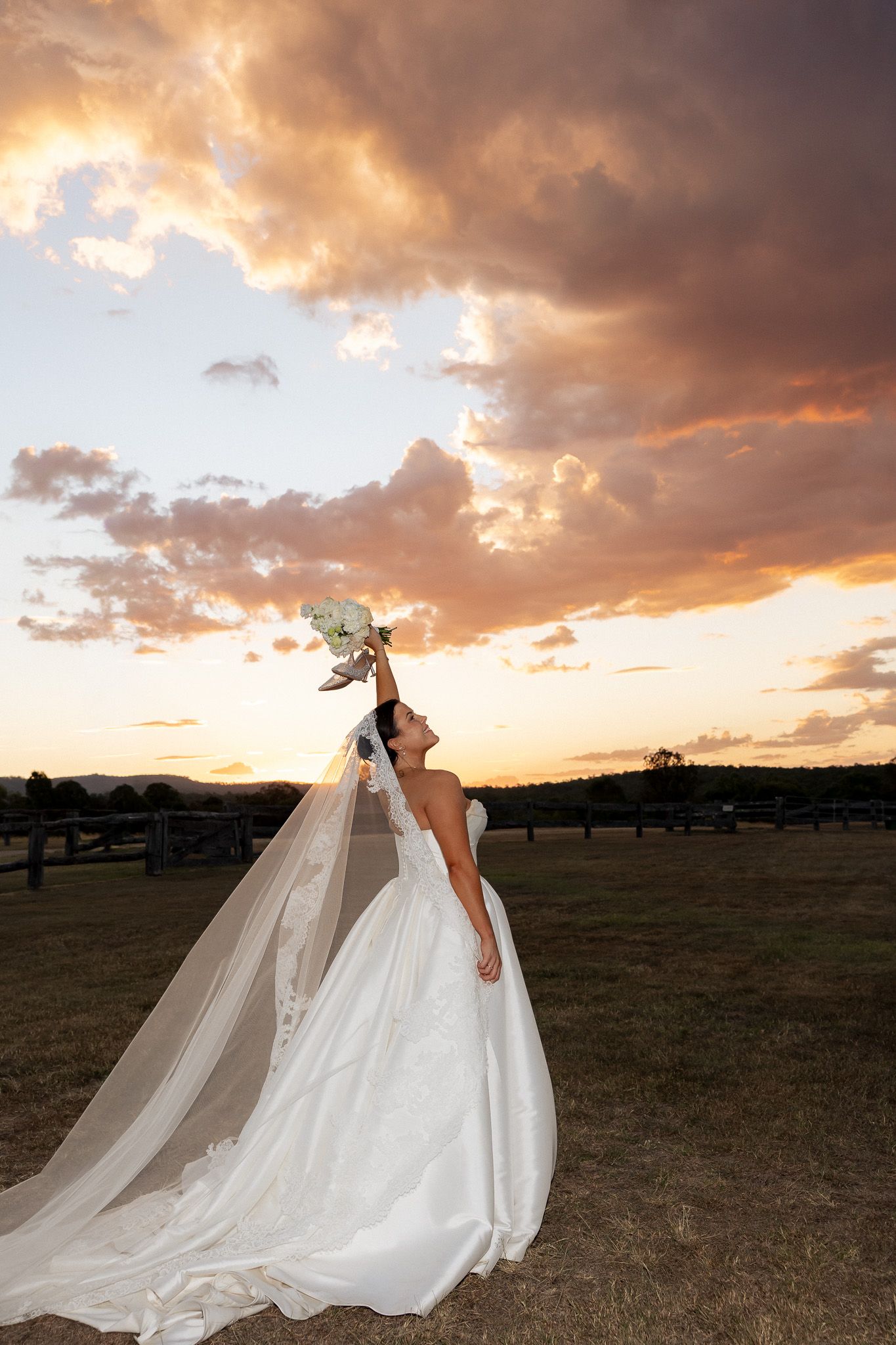 Bride in white gown raises bouquet toward dramatic sunset sky with flowing veil in rural setting.