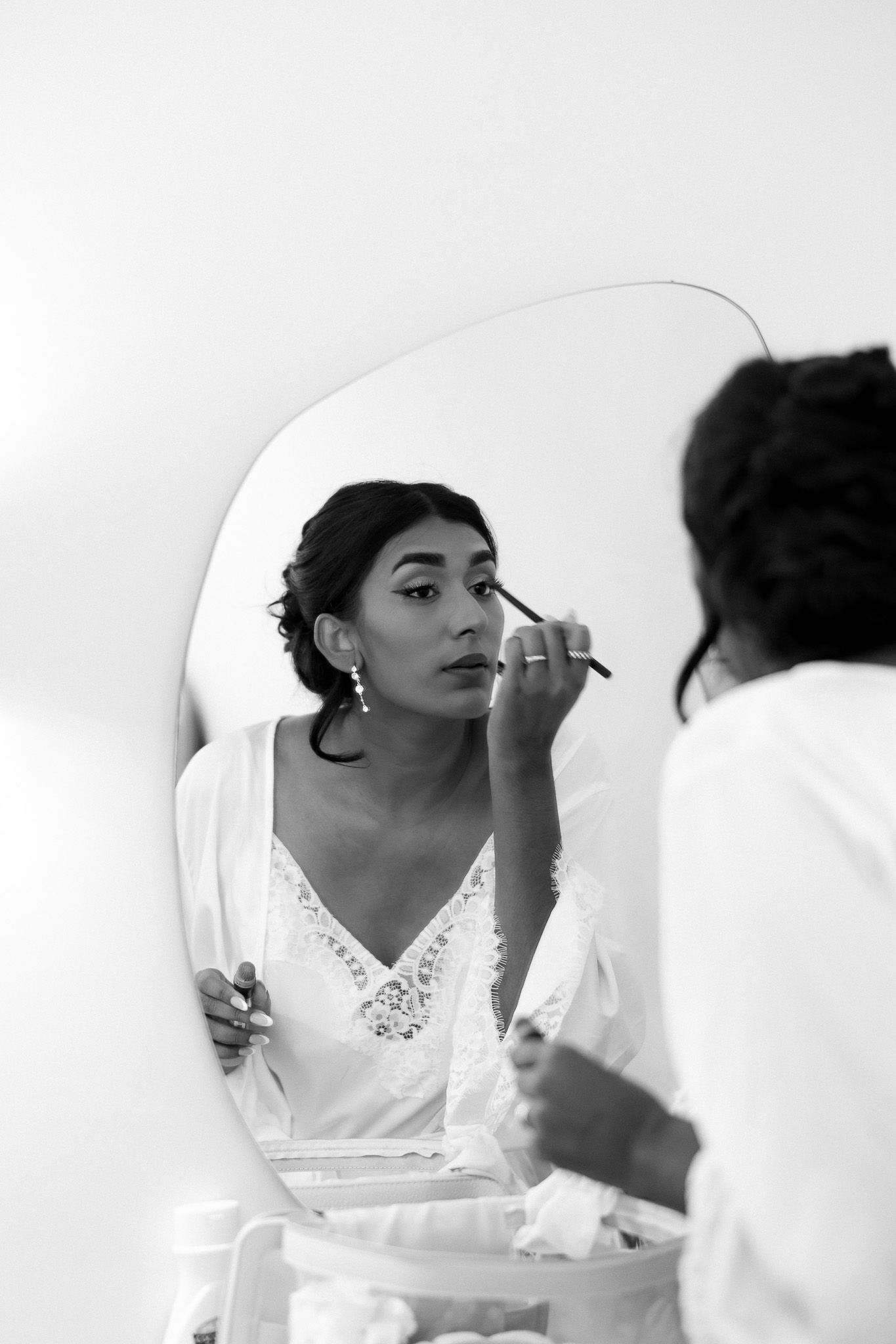 Black and white photo of bride getting makeup applied while looking in mirror on wedding day.