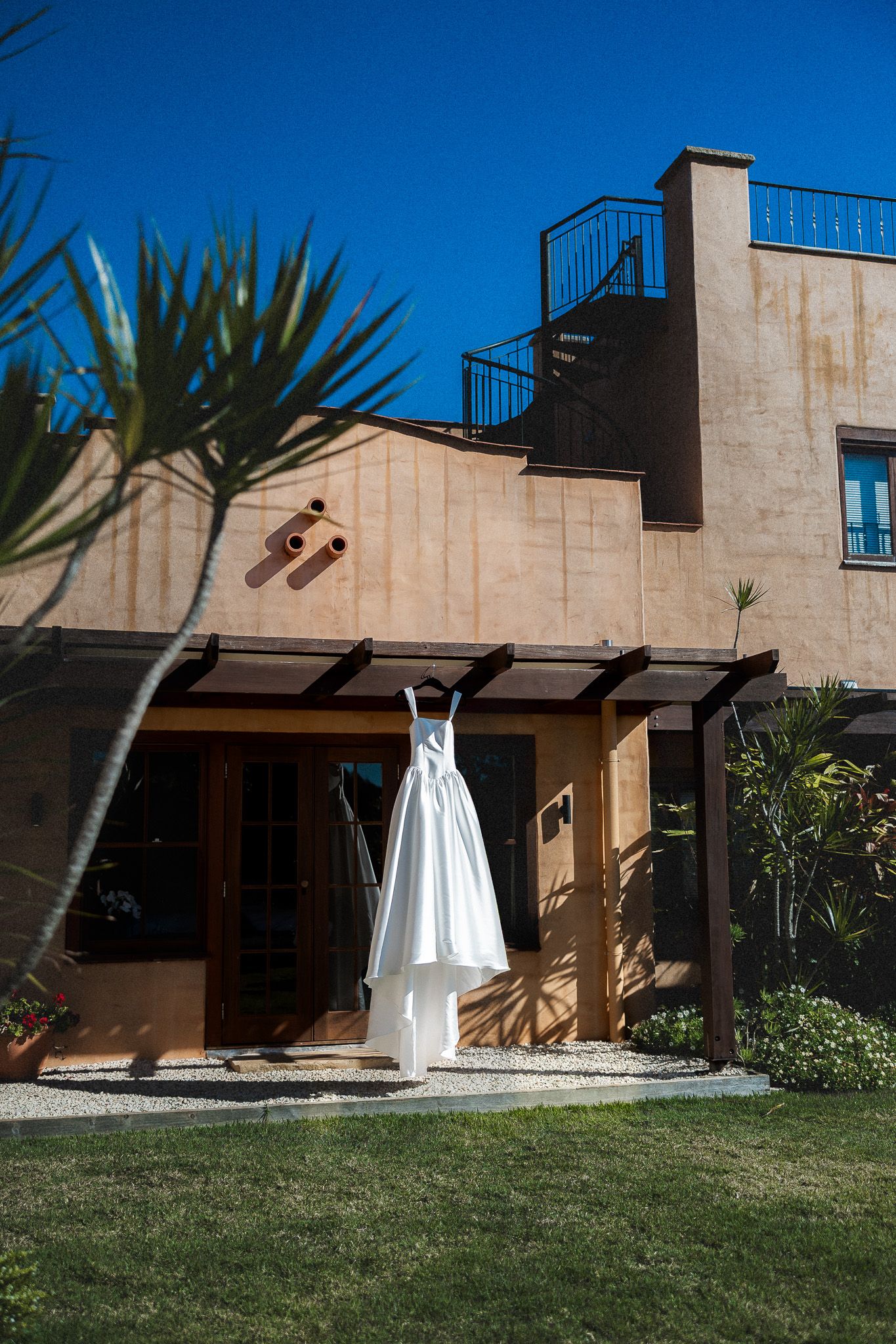 White wedding dress hanging on pergola outside rustic venue under bright blue sky.